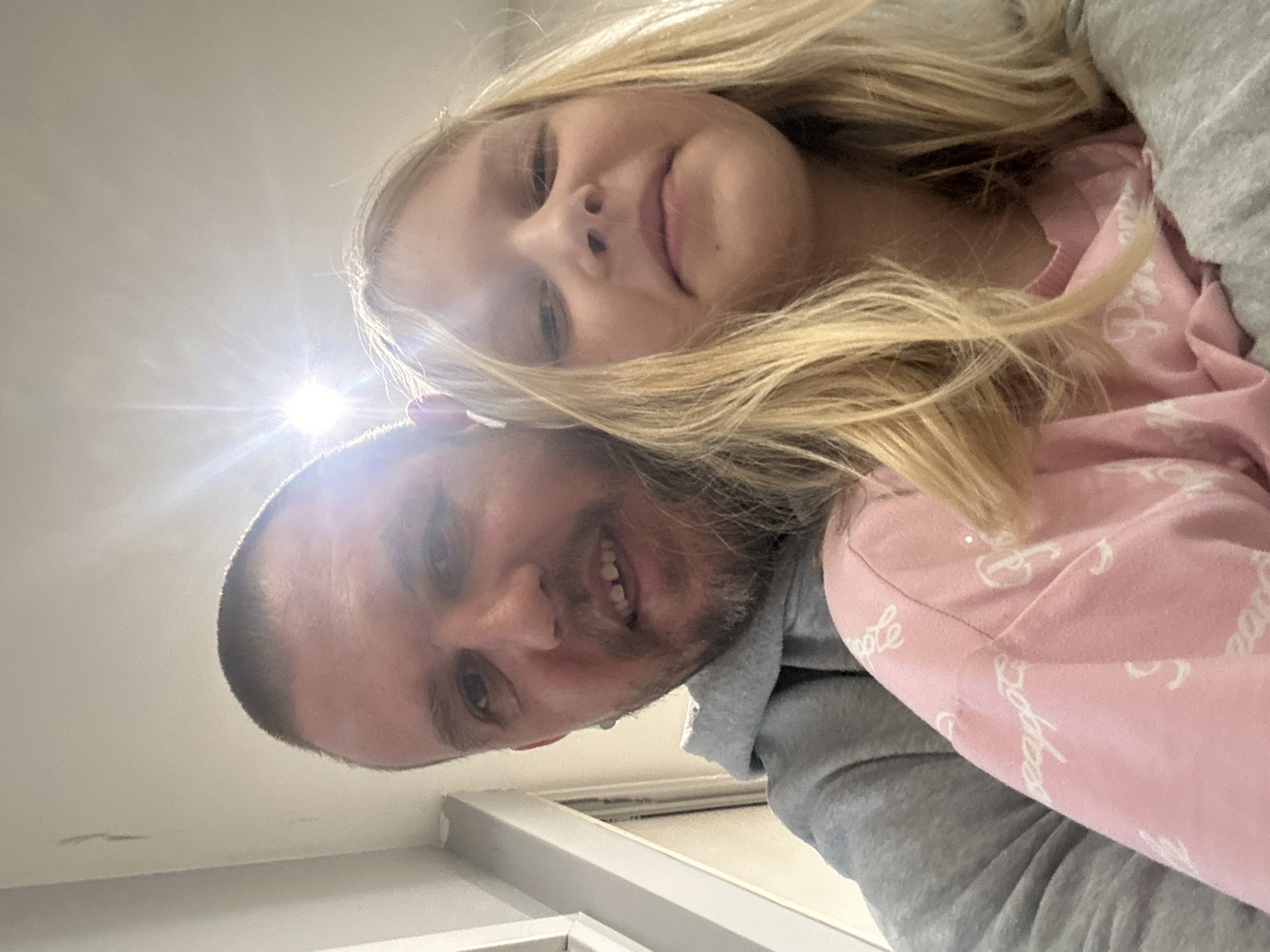 A low-angle selfie shows a man and a young girl. The girl has blonde hair and is wearing a pink top with white writing on it. The man has short hair and a light beard. He is wearing a grey hoodie. The background includes a white ceiling and a window frame. The city and country are not ascertainable from the image.