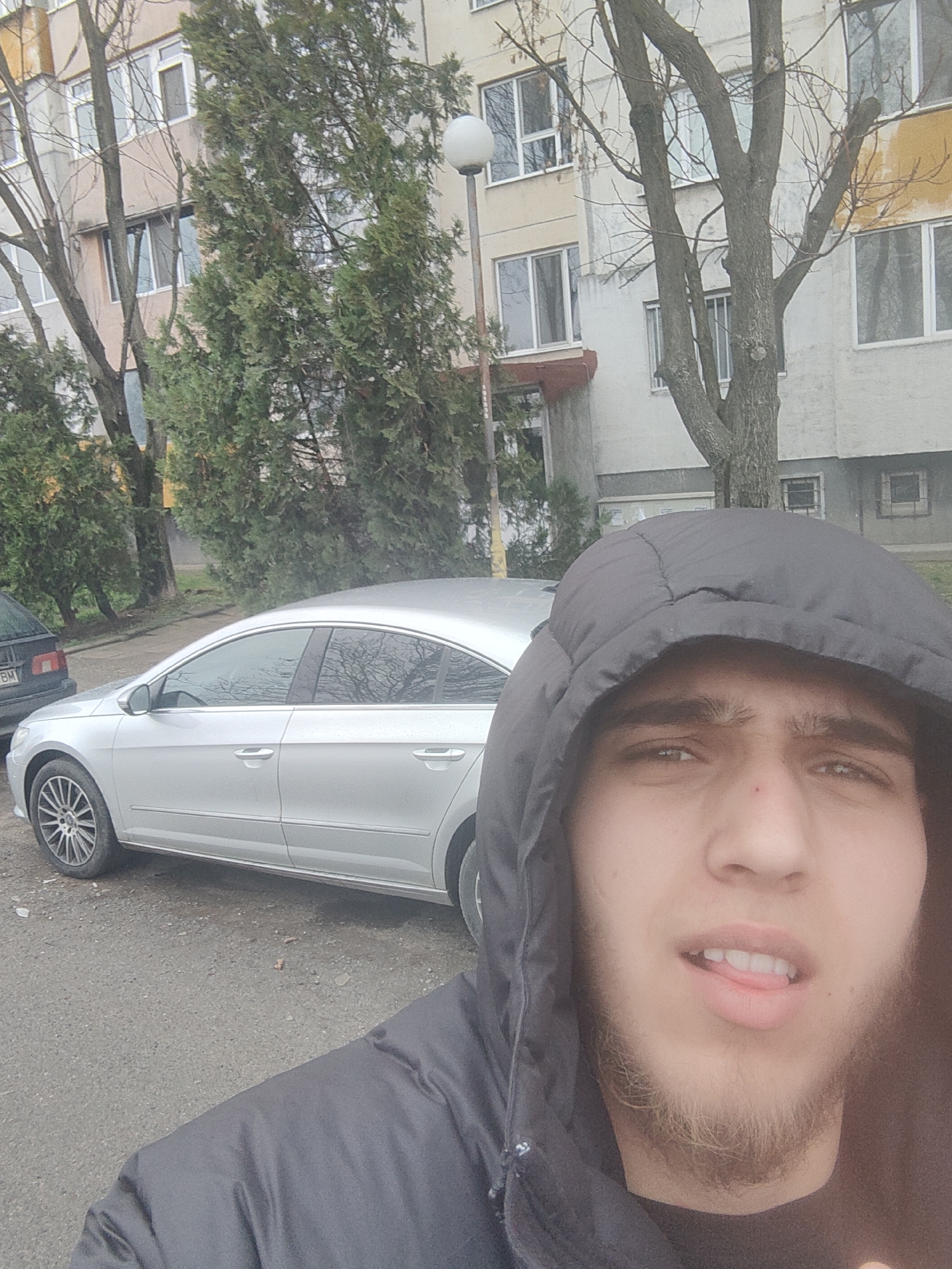 A selfie of a young man with a goatee, wearing a black hooded jacket, with his tongue slightly extended. Behind him, a silver car is parked on a paved area next to a multi-story building with trees nearby. Part of another car is visible to the left. The general setting suggests an urban environment. There is no explicit indication of the location in Sofia, Bulgaria, but the scene is suggestive of a residential area.
