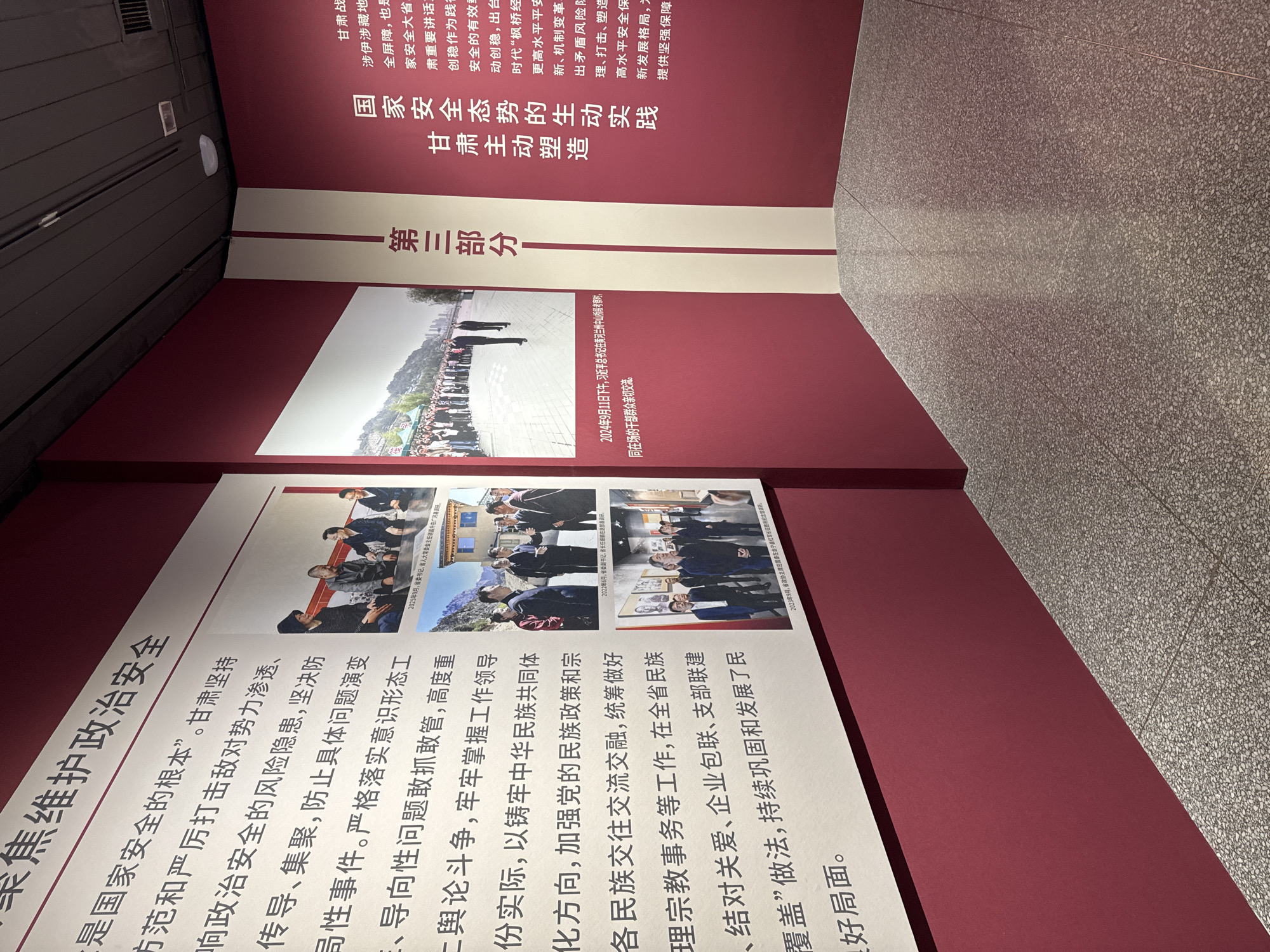 The image shows an indoor exhibition in the West Station Subdistrict, China, featuring displays about national security and political stability. The exhibits are presented on burgundy panels with white stripes, showcasing text and photographs in Chinese.
The text discusses topics such as preventing infiltration of opposing forces, preventing the evolution of issues into bigger events, and upholding the unity of the Chinese nation. The photographs depict various government officials conducting research and engaging with the public in different locations within the province.