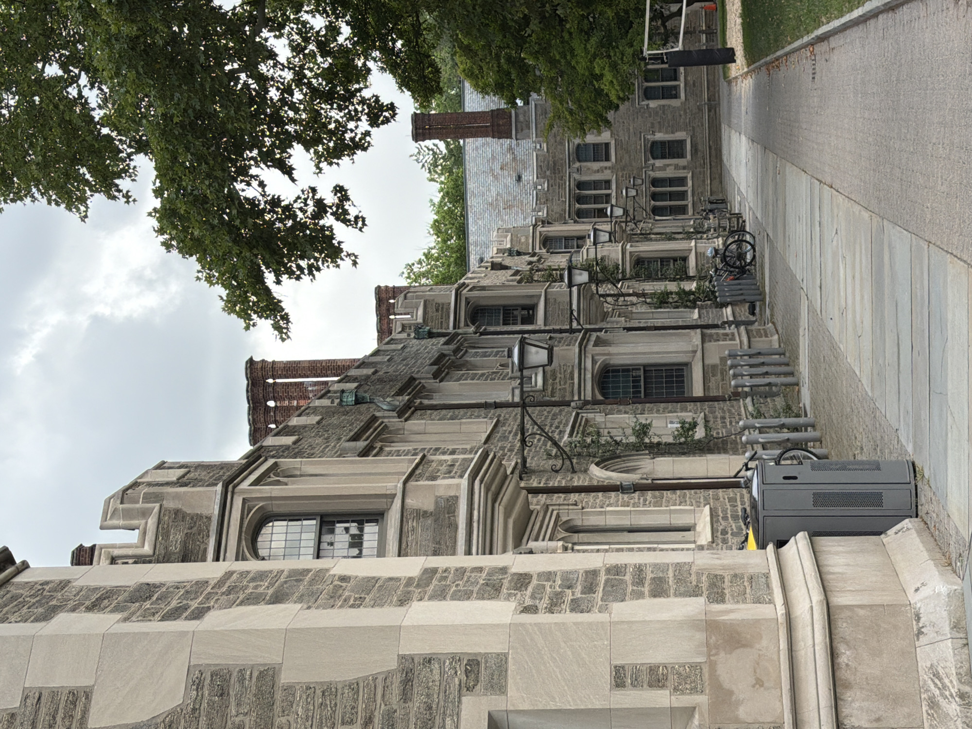 The image depicts a section of a university campus in Princeton, United States of America. In the foreground, a paved walkway runs alongside a large, stone building with Gothic architectural elements, including arched windows and decorative stonework. The building appears to be multiple stories high, constructed from a combination of rough-hewn gray stone and lighter-colored carved stone. Several wrought-iron lampposts are attached to the building's facade.
Further down the walkway, a bicycle is parked near a series of metal bollards. Adjacent to the walkway, a gray, rectangular trash receptacle is visible. Beyond the building, a line of trees with dense green foliage creates a natural backdrop, with some branches extending into the upper portion of the frame. The sky is overcast, suggesting a cloudy day. In the distance, another building with a similar stone construction and peaked rooftops is partially visible. A volleyball net and post are also present on the right side of the image, indicating recreational facilities. The scene is devoid of any people.