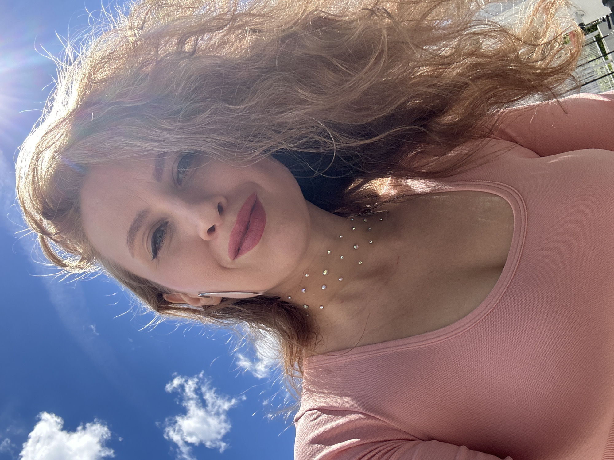 A woman with blonde, wavy hair and pink lipstick is taking a selfie outdoors. She is wearing a pink, long-sleeved shirt. The sky in the background is blue with white clouds, and the sun is shining brightly, creating a lens flare. She has a necklace made of small, shiny stones around her neck and is wearing a long, silver earring. The image appears to be taken from a low angle, looking up at the woman and the sky. There are no indications of Krasnogorsk, Russia in the image.