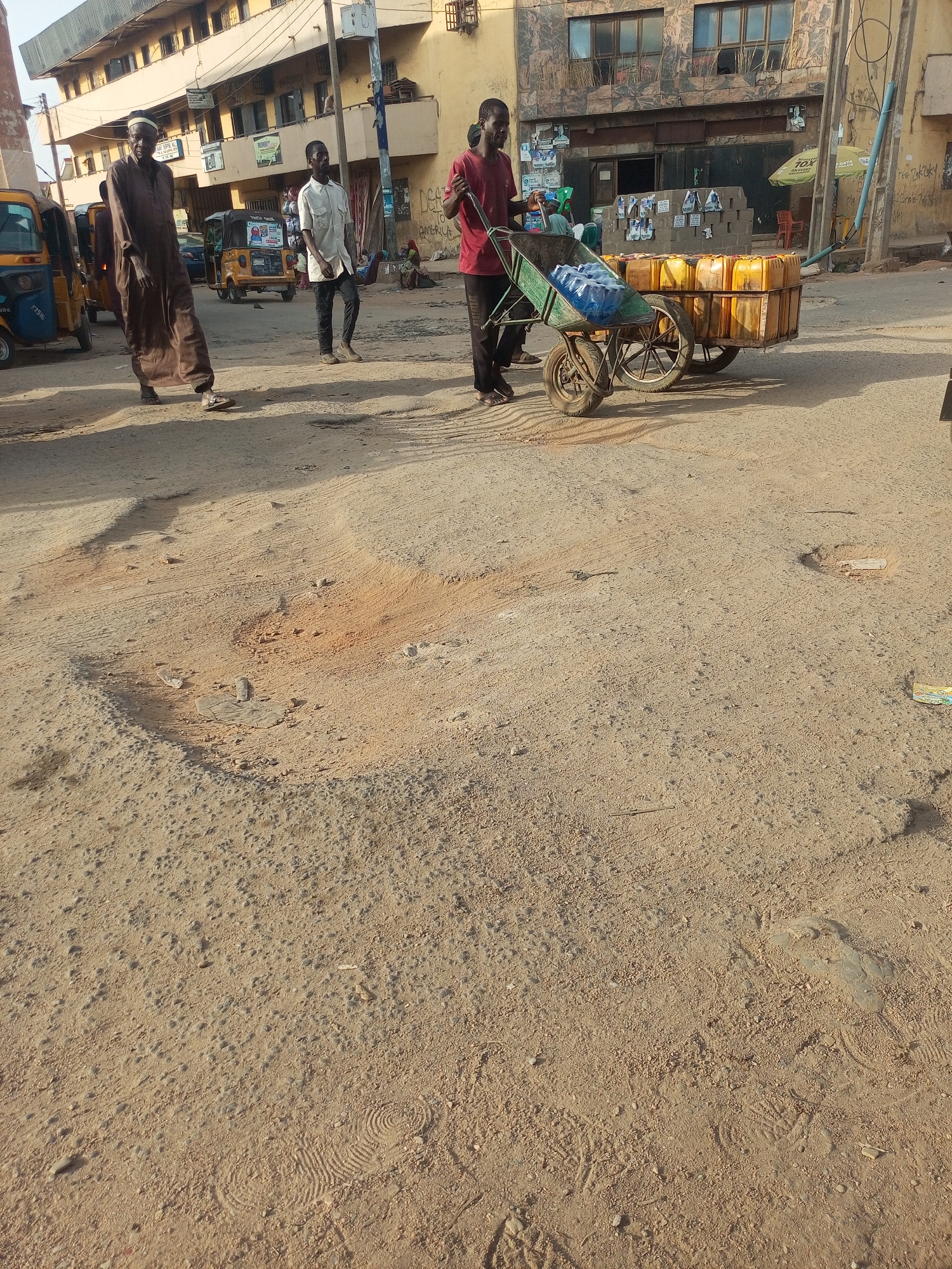 The image depicts a street scene in Maiburiji, Nigeria. Three men are visible: One, wearing a long brown robe, walks left. Another, in a light-colored shirt and dark pants, walks to the right. A third man in a red shirt pulls a two-wheeled cart loaded with blue water bags and yellow jerrycans. Behind him is another cart loaded with yellow jerrycans. The street is unpaved with visible potholes and footprint impressions. A row of auto rickshaws is parked on the left side of the frame. Buildings line the street, displaying a mix of residential and commercial use. Graffiti can be seen on one of the buildings.