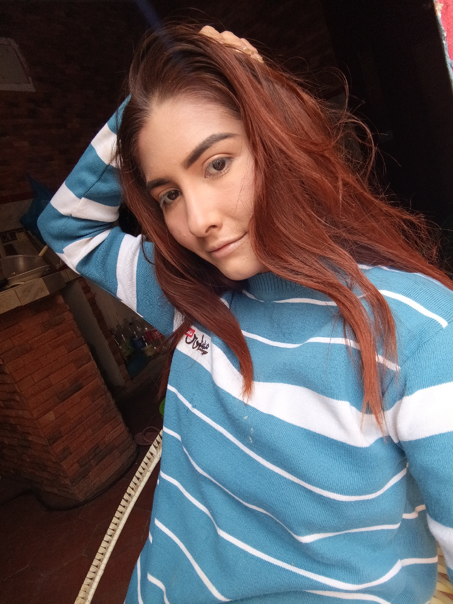 A young woman with auburn hair, wearing a blue and white striped sweater, poses for a selfie. Her right hand rests on her head, and she is gazing downwards with a slight smile. The background is a blend of indoor and outdoor elements, featuring a brick wall and a dark, undefined area that could be a doorway or window. The presence of what seems to be a cooking space nearby further indicates the setting as a home or indoor area. The image was likely taken in Juan Griego, Venezuela.