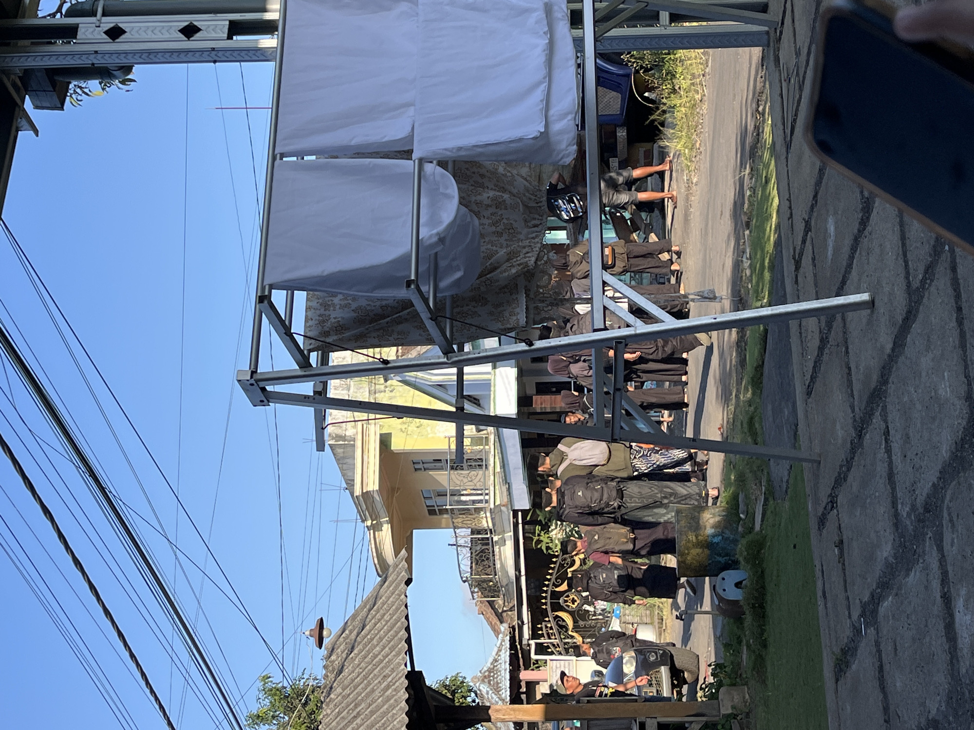 In the city of Jambearum, Indonesia, a group of individuals, most wearing dark clothing and head coverings, are gathered outdoors on a paved area adjacent to a street. The scene is illuminated by sunlight, indicating it is daytime. A collection of overhead power lines crisscross the bright blue sky. In the foreground, a metal A-frame structure holds several white fabric sheets, possibly being used for drying or display, along with patterned fabric panels behind them. A dark-colored car is parked to the left, and behind the group, buildings with balconies and decorative railings are visible. A small, blue and yellow cylindrical object with a face painted on it sits on the grass to the left of the group. The ground is a mix of concrete paving slabs with visible lines and patches of grass. The presence of a mobile phone in the bottom right corner suggests someone is documenting the scene.