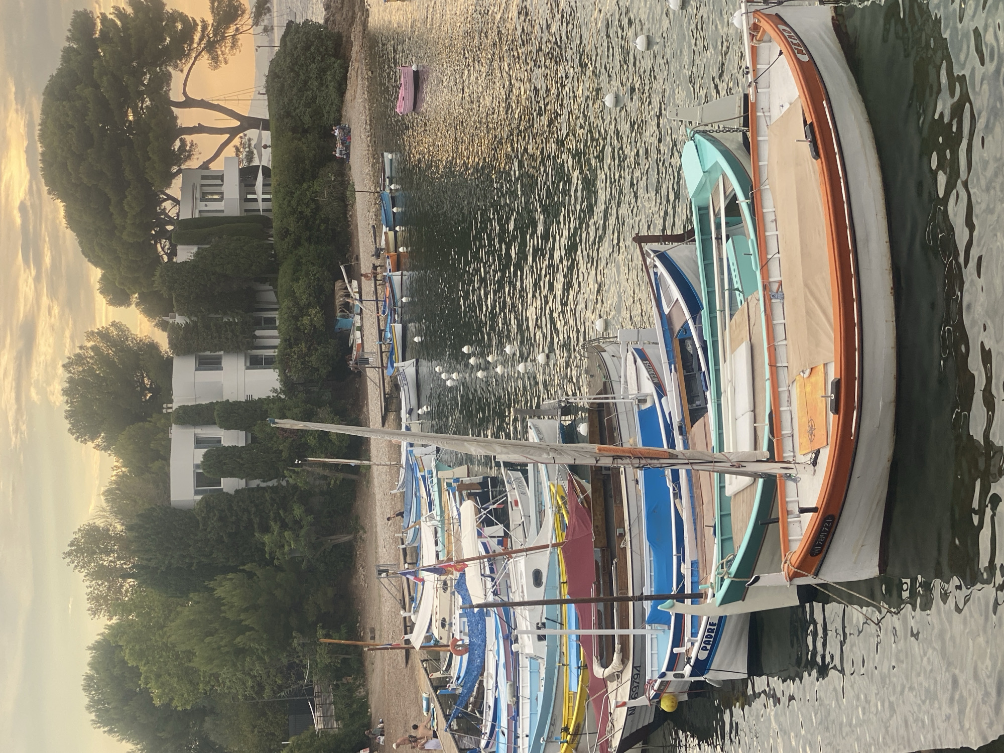 The image depicts a harbor scene in Antibes, France, at what appears to be sunset. Numerous small boats are moored in the foreground, lined up along a dock. The boats are of various colors and designs, with some featuring sails and others equipped with outboard motors. Several boats have covers over their seating areas, and some are marked with registration numbers and names, such as "PADRE" and "NEMO."
In the middle ground, a gravel beach is visible, with a few people seated at tables, possibly dining or socializing. Behind the beach, a modern white building with large windows is nestled among lush green trees. Further in the background, more trees and foliage line the coast, with glimpses of sailboats in the water. The sky above is a soft yellow and orange, indicative of a sunset, with scattered clouds. The water is calm, reflecting the warm hues of the sky and the surrounding environment. The overall impression is one of a peaceful, scenic waterfront location.