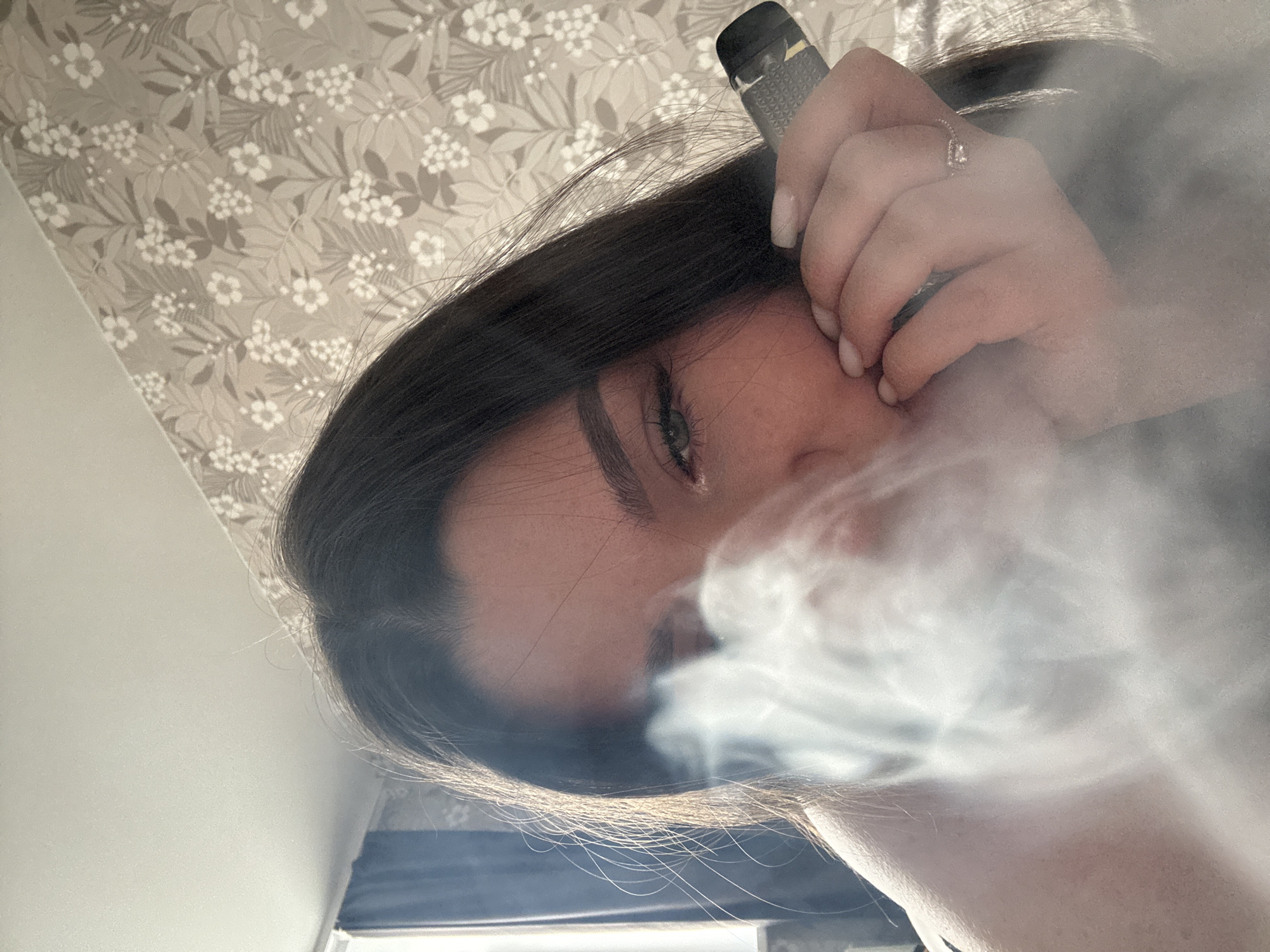 A woman is depicted at an upward angle, blowing out a cloud of smoke. She is holding a vape pen. She has dark hair, an eyebrow piercing, and is wearing a ring on her right hand. The background includes a floral wallpaper pattern. The presence of Russian city or environment elements cannot be ascertained from the visual information of this image.