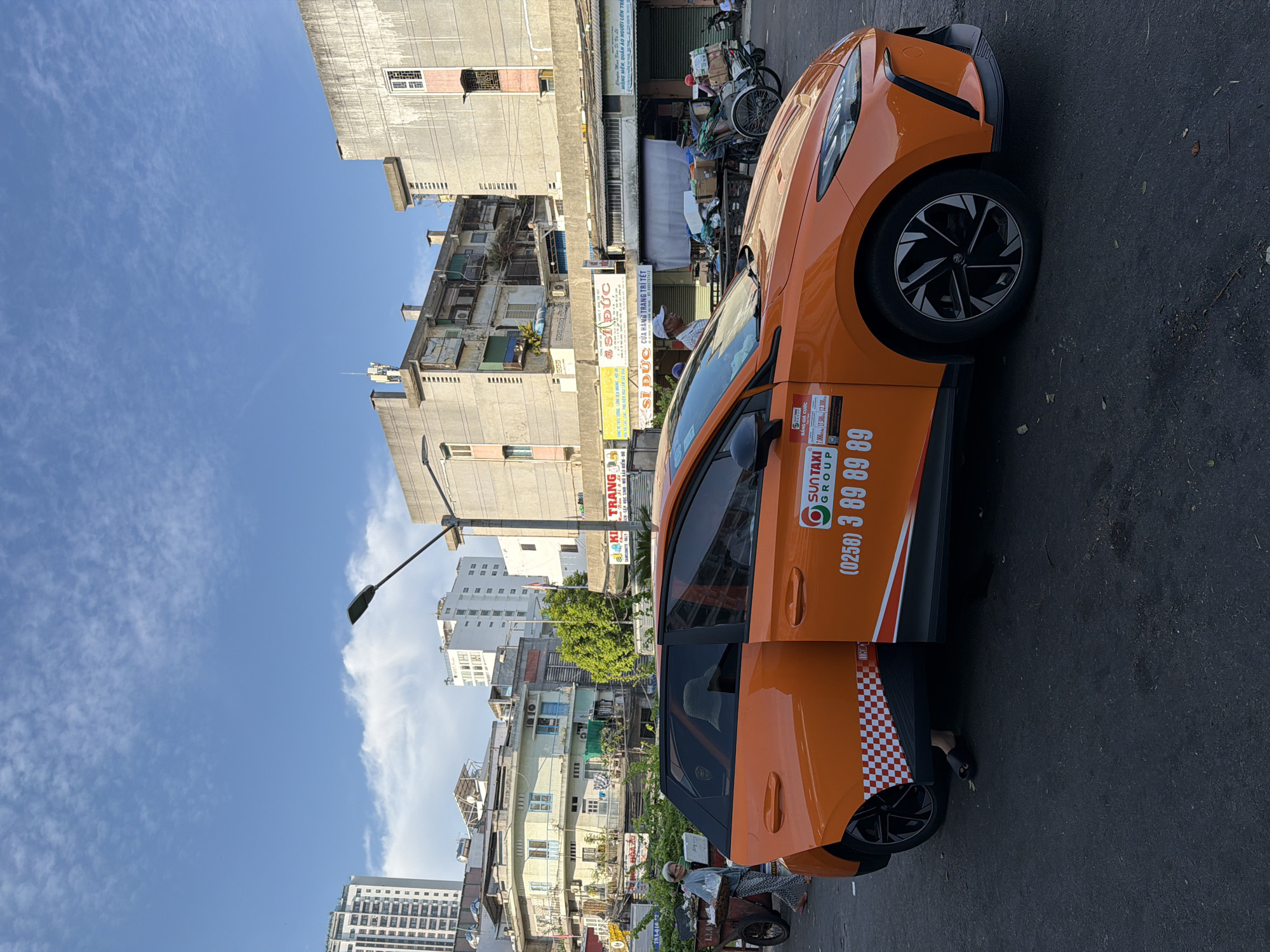 In Nha Trang, Vietnam, an orange taxi with its front passenger door ajar is parked on an asphalt road. The taxi displays "SUNTAXI GROUP" and the phone number "(0258) 3 89 89 89" on its side. To the left of the taxi, an elderly woman wearing a light blue shirt and patterned pants walks past a handcart laden with items, while another person, possibly a passenger, is visible inside the taxi wearing a straw hat. In the background, a bustling urban scene unfolds with buildings of varying heights, shops with open fronts, streetlights, and a blue sky with scattered clouds. A man in a white cap is seen near the shops, and a bicycle rickshaw loaded with goods is also present. The overall environment suggests a commercial area or marketplace in the city.