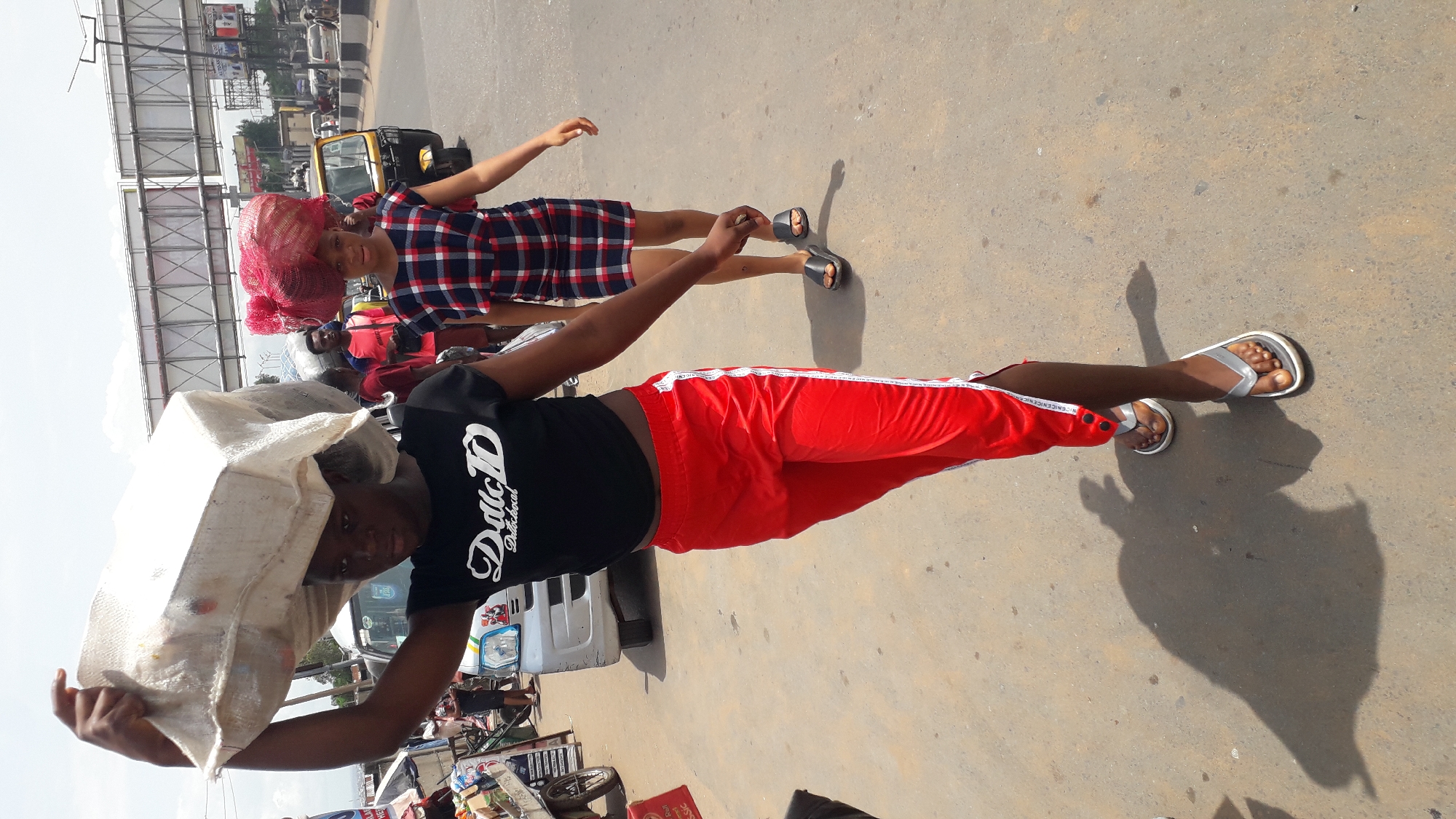 The image depicts two people in what appears to be a street or roadside setting. One person is carrying a large sack on their head, wearing a black T-shirt with white lettering, red pants with a white stripe, and sandals. The other person is wearing a red hair covering, a plaid dress, and sandals. The background shows vehicles, including a yellow auto rickshaw and other parked cars, suggesting an urban environment. The image is presumably taken in Onitsha, Nigeria, given the street and type of vehicles in the background. The surface beneath their feet appears to be paved.