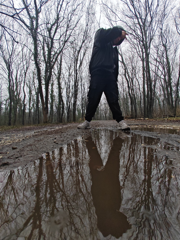 An individual stands centrally on a muddy, unpaved path within a forest located in or adjacent to Shumen, Bulgaria. The subject, an adult, wears a dark hooded jacket or top, dark cargo-style trousers, and light-colored athletic shoes. The person's head is lowered, and both arms are raised and bent, with hands positioned near the head/face. The legs are spread in a wide, stable stance. A substantial puddle of water occupies the foreground and mid-ground of the path, reflecting the bare, intricate branches of numerous leafless deciduous trees that line both sides of the path. Small concentric ripples are visible on the puddle's surface. The sky is overcast, and the ground is consistently wet and muddy. The absence of foliage on the trees indicates a late autumn or winter season. No other persons or specific man-made objects are present in the frame.