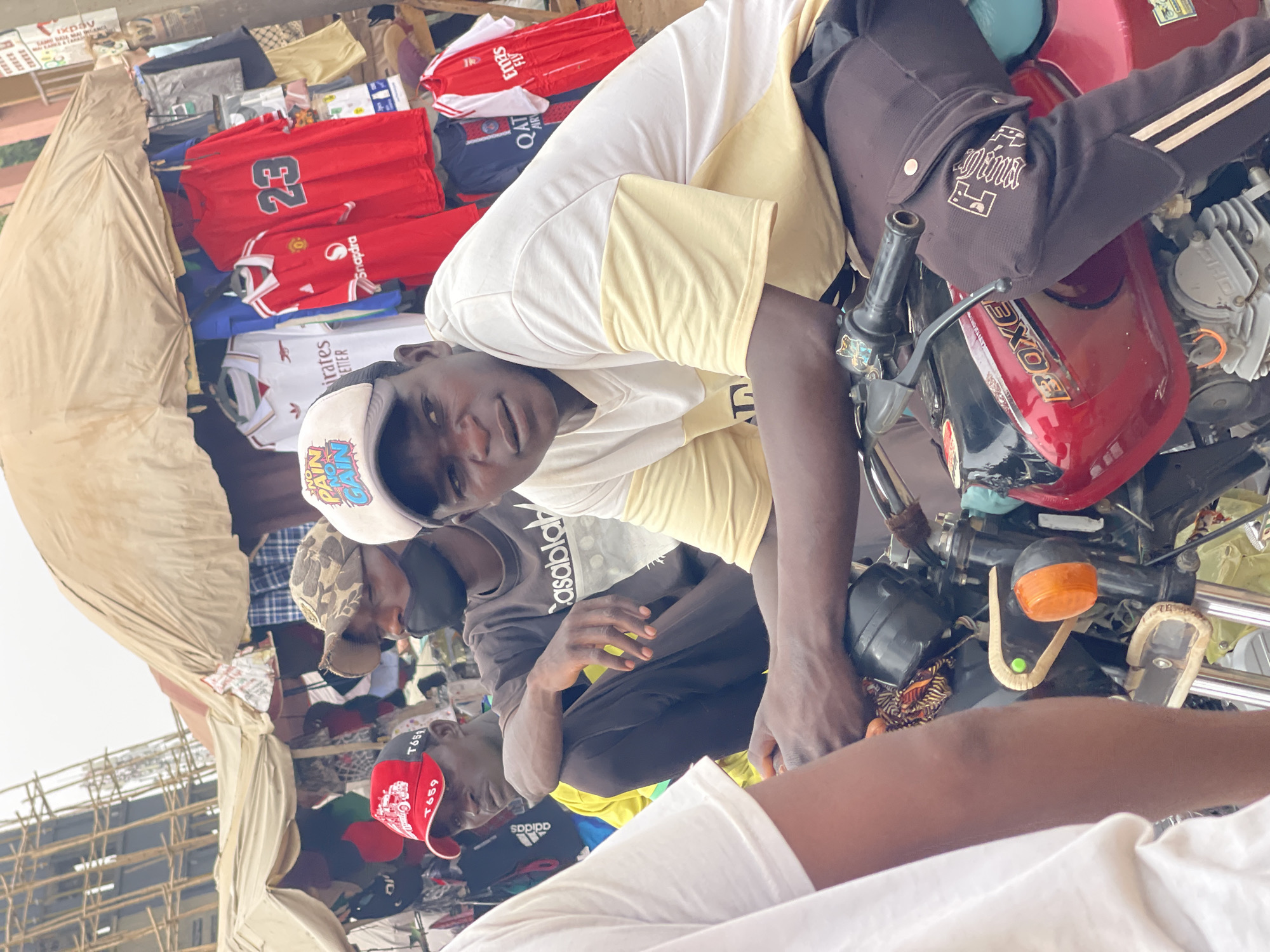 This image, set in Chikaji, Nigeria, captures a bustling street or market scene during daytime. The primary focus is on several men gathered around a red motorcycle and a clothing stall.
In the foreground, a young man looks directly at the camera. He wears a white and yellow short-sleeved shirt and a white baseball cap with colorful text that reads "NO PAIN GAIN". His hands are positioned near the handlebars of a prominent red motorcycle. The motorcycle, branded "BOXE" on its fuel tank, has black handlebars and an orange indicator light. A dark, possibly black, jacket with light-colored stripes and text resembling "Dina" is draped over the motorcycle's front.
Seated or stooping behind the main subject, on the motorcycle, is another man wearing a black long-sleeved top with "Casablak" (likely "Casablanca") written on it, and a light-colored, possibly camouflage, cap. Further to the left and rear, a third man is visible, wearing a red and black cap with "T659" printed on it. Part of a fourth person in a white shirt is seen in the bottom right corner.
To the left of the men, an informal clothing stall operates under a light-colored canopy. Numerous football jerseys are displayed, including red Manchester United shirts (one with "23" and "Snapdra" logos), a white Arsenal "Emirates" jersey, and a dark blue Paris Saint-Germain (PSG) shirt with "QATAR AIRWAYS" visible. Other clothing items are stacked or hung.
In the background, a structure that appears to be scaffolding or a similar framework is visible. The bright lighting suggests a clear, possibly sunny day. Other visible text includes "SDPIPD", "ixpay", "FLY", and "Ména" on various items in the scene. The men appear to be engaged in a casual interaction, possibly related to selling or purchasing items at the stall.