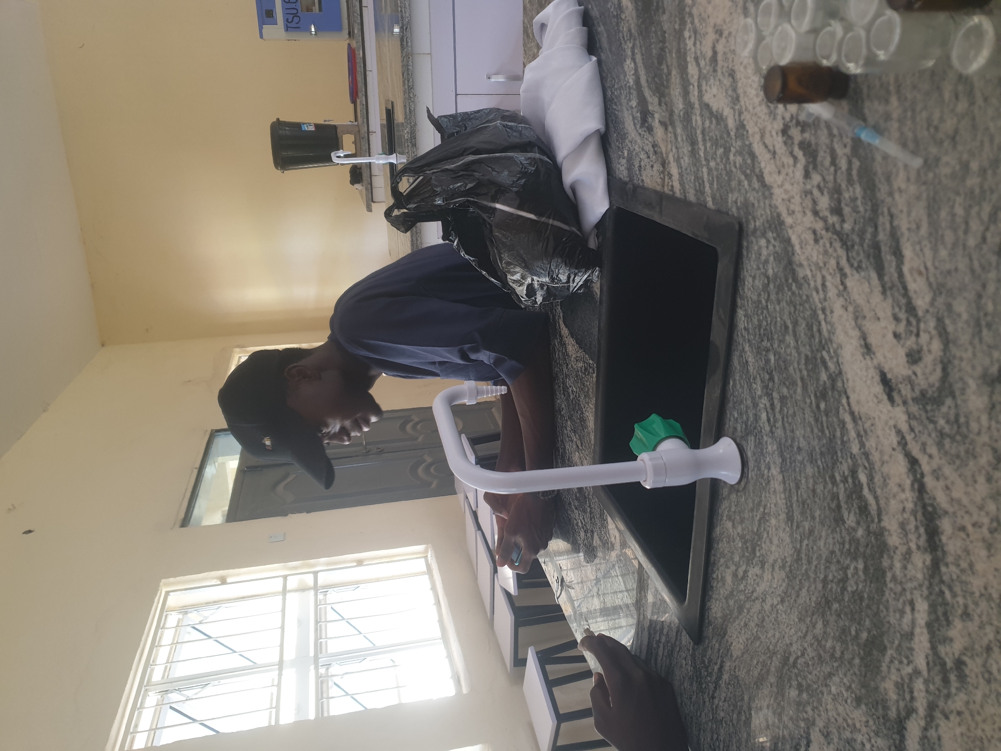 This image shows a person wearing a dark blue shirt and a black baseball cap, looking down at their phone, seated at a laboratory counter in Jalingo, Nigeria. To the left of the person, another individual's hand is visible, also appearing to be holding a phone. The counter is made of speckled granite, and a white sink with a green-handled faucet is prominent in the foreground. Behind the sink, a black plastic bag and a white cloth are placed on the counter. In the background, a window with metal bars overlooks an outdoor area, and several white stools are lined up. A laboratory sink, a water dispenser, and a blue and white piece of equipment are visible further back on the counter. The walls are painted a light yellow, and a door is visible to the left. Small vials and a syringe are on the counter to the right, indicating a scientific or medical setting.