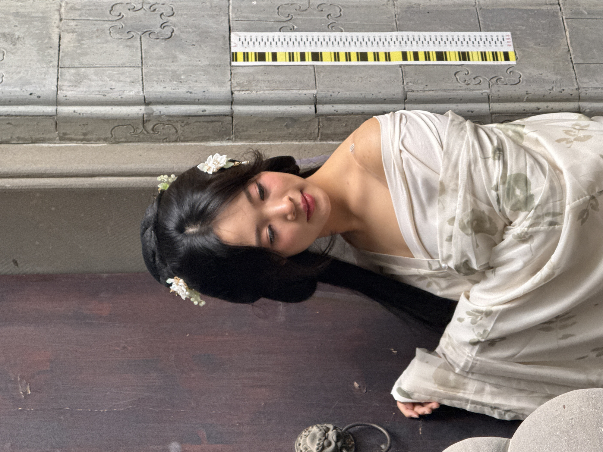 A woman is reclining on a dark brown wooden surface, presumed to be a floor, with her head near the left edge of the image. She is wearing a light-colored robe with a floral pattern. Her dark hair is adorned with small white flowers, and she is facing the viewer with her eyes closed. Behind her is a light gray wall with decorative cloud-like patterns. At the top of the wall, a measuring scale with black and yellow markings is affixed. A decorative metal object is located near the bottom left. The scene is consistent with architectural details found in Yuzhong District, China.