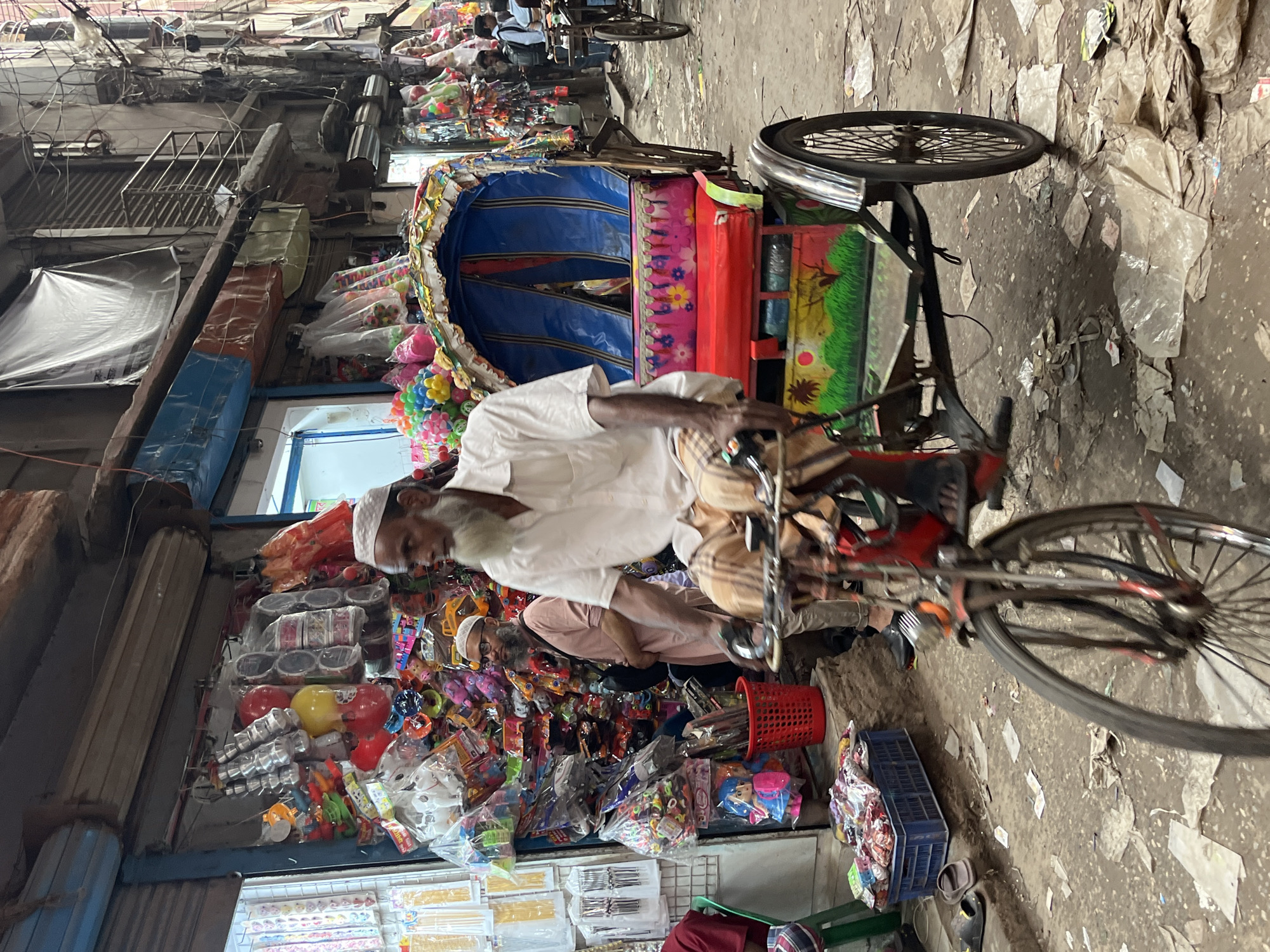 The image portrays a busy street scene in Dhaka, Bangladesh. A man, likely in his 60s or 70s and wearing traditional Muslim attire with a white skullcap, is riding a colorfully decorated cycle rickshaw. The rickshaw's passenger area features a blue and red canopy. The street is cluttered with various goods for sale, including toys, balloons, and small plastic items, indicating a market area. The buildings on the left side are old and have multiple levels, with awnings and metal railings visible. The ground is covered in litter and debris. There are other pedestrians in the background, and another bicycle is partially visible. The image is taken from a slightly elevated perspective.