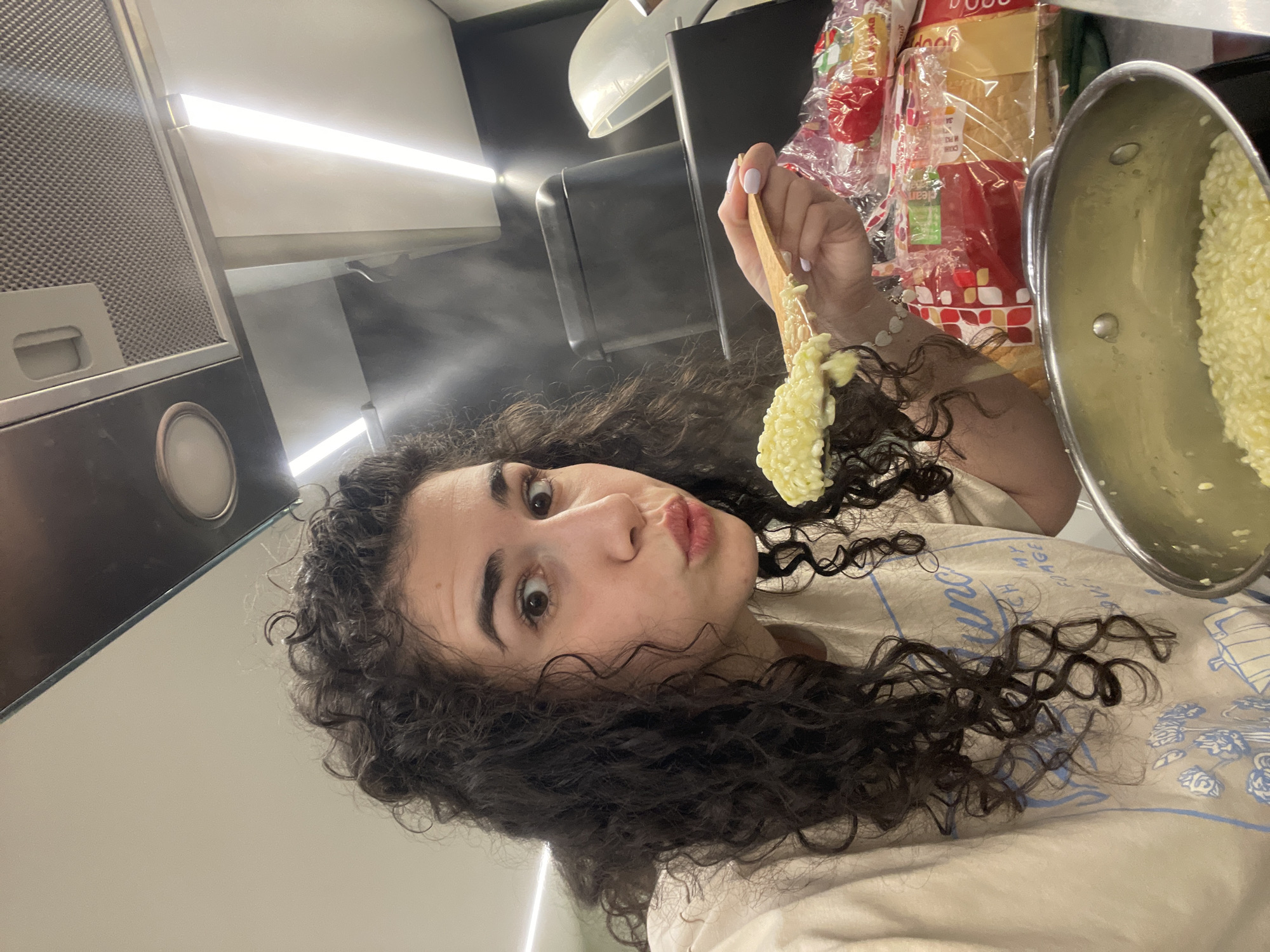 A person with curly dark hair is in a kitchen. They are holding a wooden spoon with cooked rice in one hand, and a pot of rice in the other hand. The background includes a kitchen hood, a light fixture, a loaf of bread, and other kitchen items. There is steam rising from the food. The location of the photo is not evident from the provided image; therefore, the location in Sofia, Bulgaria is not determinable.