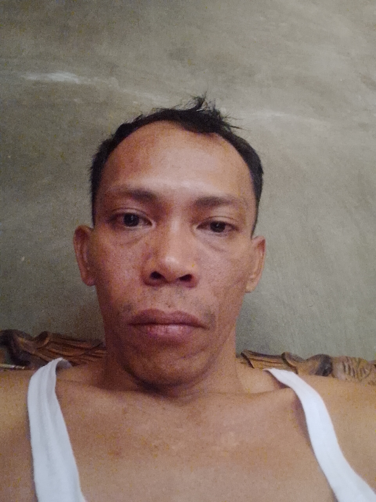 A medium shot shows a close-up of a man's face and upper chest. The man has dark hair, dark eyes, a wide nose, and thin lips. He is wearing a white tank top. The background is a textured, gray wall. The scene appears to be indoors, possibly in a home, as indicated by the presence of what appears to be furniture or a bed behind the man. The lighting is natural and appears to be coming from the front. There is no specific information to determine the location in the city of Madukoro, Indonesia, from the visual evidence alone.
