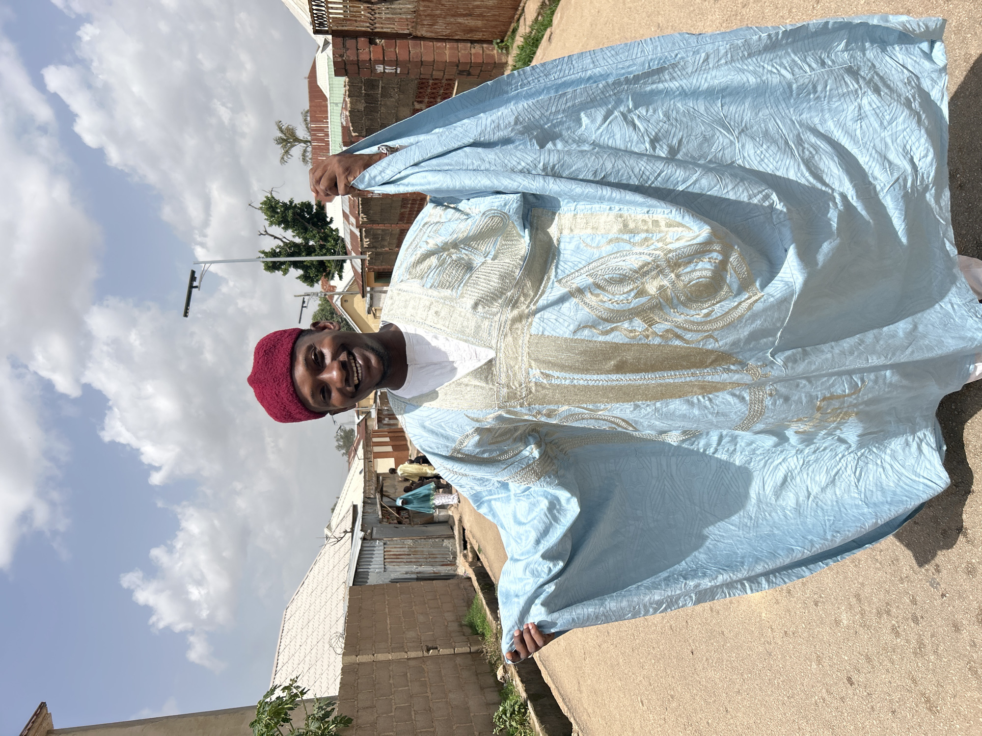 A man with dark skin and a red fez smiles at the camera, holding a light blue boubou garment with cream-colored embroidery. He stands on a light-colored concrete surface, likely a sidewalk or street. Behind him, there's a neighborhood scene with buildings constructed from brick and corrugated metal, typical of Jos, Nigeria. Utility poles and wires are visible in the background sky. The sky is partially cloudy.