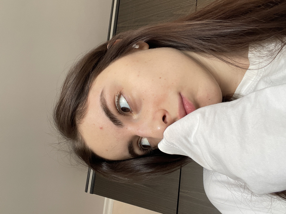 A young woman with long, dark brown hair is pictured leaning against a white, soft-looking object. She's looking upwards with her brown eyes. Her skin is fair with visible freckles and subtle makeup. The background is a neutral-toned wall with a dark wooden door or cabinet visible in the frame. The image is slightly rotated.