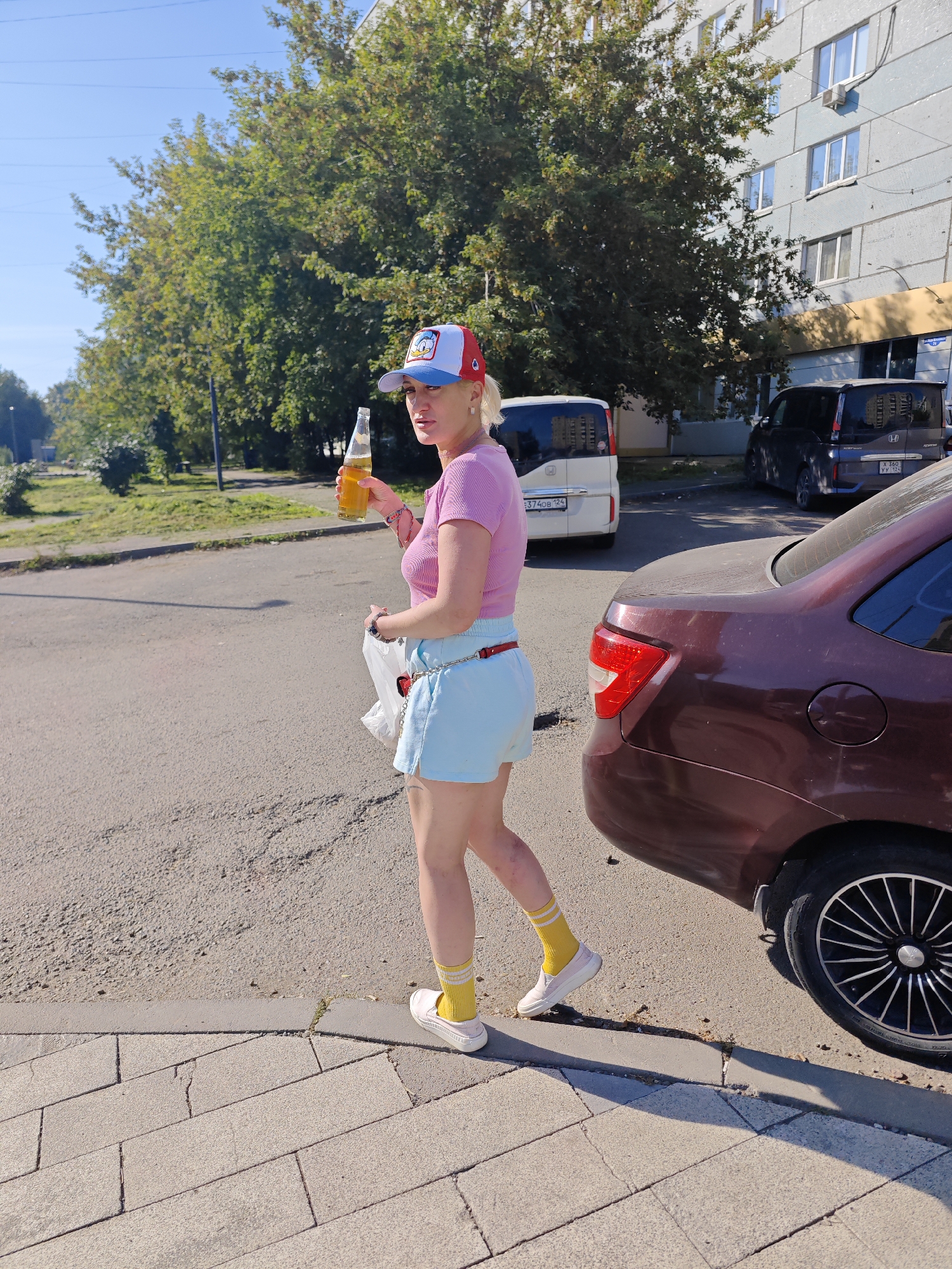 A woman stands on a sidewalk next to a maroon sedan in a residential area. She is wearing a pink t-shirt, light blue shorts, yellow socks, and white shoes, and a red, white, and blue baseball cap. She holds a bottle of amber liquid in her right hand and a white plastic bag in her left. In the background, a multi-story apartment building and trees are visible. The scene appears to be a sunny day in Krasnoyarsk, Russia.