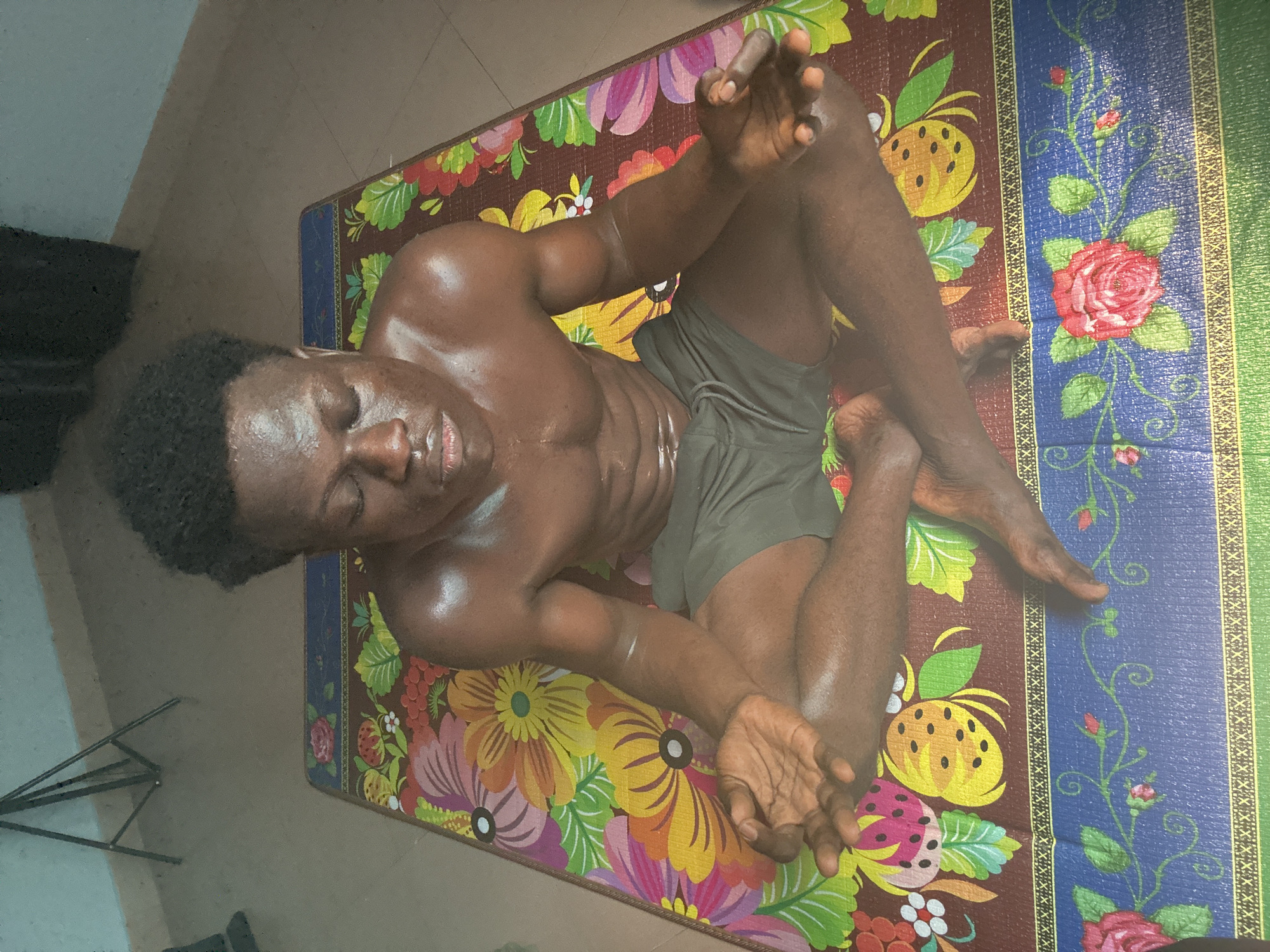 A young Black man with a muscular physique is seated in a lotus position on a colorful, floral-patterned mat. His eyes are closed, and his face appears relaxed. His hands are resting on his knees, with his palms facing upwards in a meditative gesture. The mat is placed on a tiled floor, and a tripod can be seen in the background, suggesting the image might have been taken in a home setting or a studio. The lighting suggests an indoor environment. The image is likely from Badagry, Nigeria, a city known for its historical significance and vibrant culture, where such mats are commonly used.
