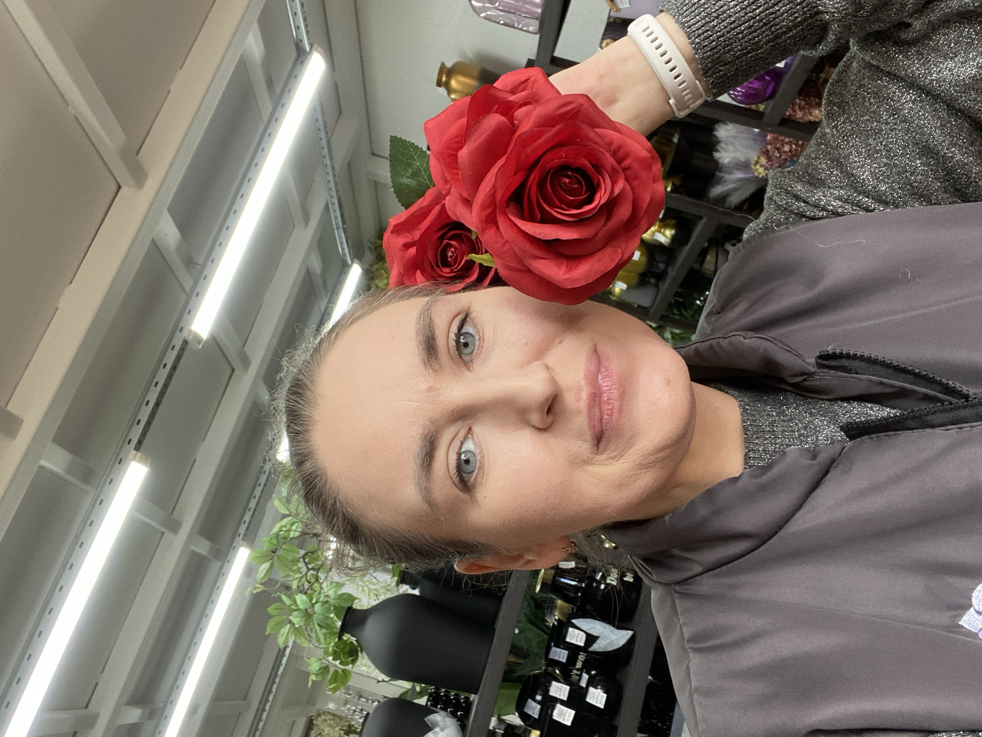 A woman holds three artificial red roses near her face. She wears a gray jacket over a silver sweater and a white smartwatch. The background shows retail shelving containing black vases, faux greenery, and other decorative items. The ceiling features fluorescent tube lighting. The location is likely a home goods store in Yekaterinburg, Russia, based on visual cues and assumptions.