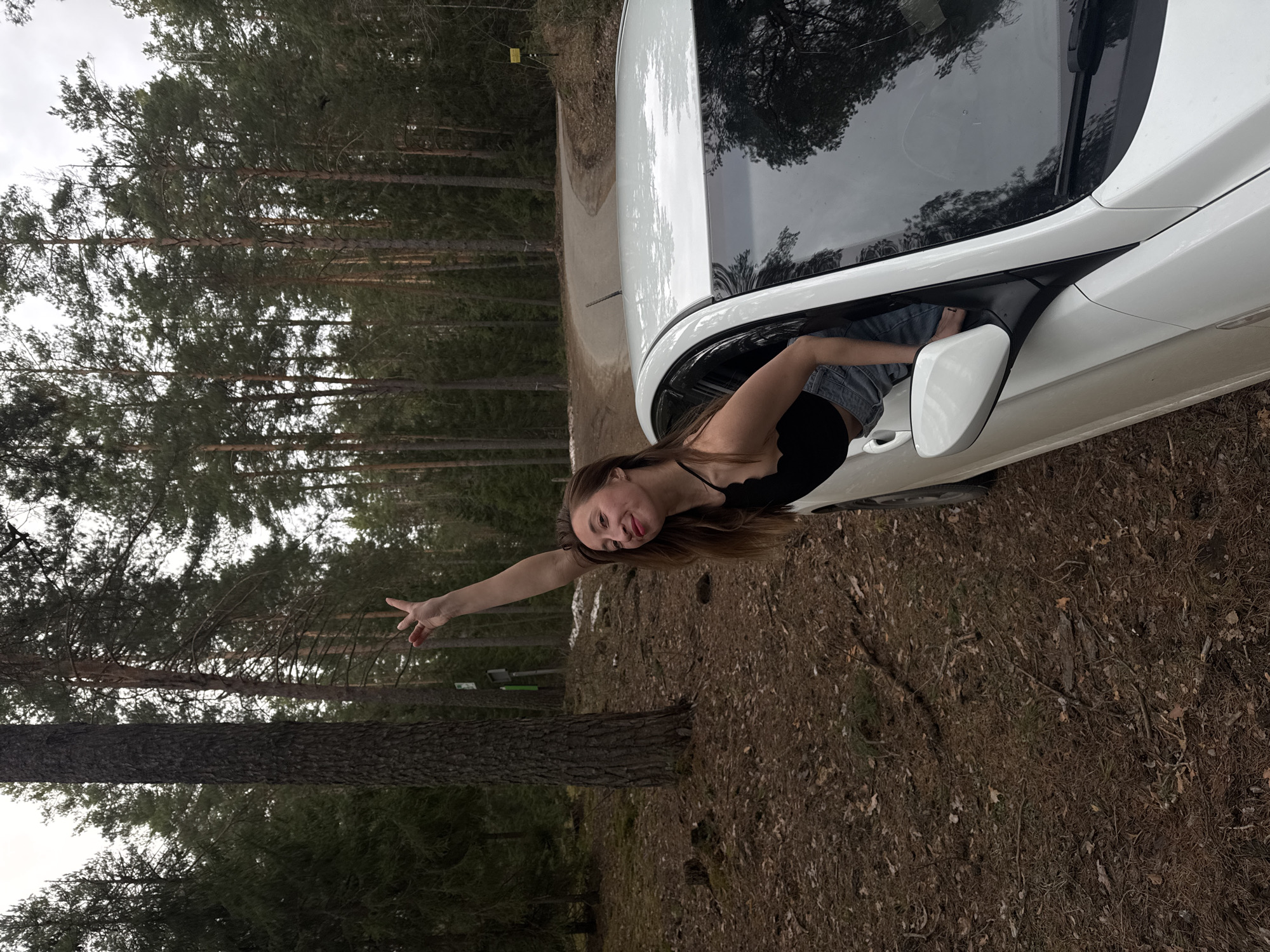 A young woman is captured leaning playfully out of the passenger side window of a white car, positioned on what appears to be a dirt track within a dense forest. She has long brown hair and is wearing a black tank top and denim shorts. A bright smile is on her face, with her tongue slightly extended, as she holds her right arm outwards, making a peace sign with her fingers.
The car, a white sedan, reflects the surrounding trees in its windows. The setting is a forest dominated by numerous tall, slender pine or conifer trees with dark, textured bark and green foliage, extending upwards towards an overcast sky. The ground is covered with a thick layer of brown pine needles, dirt, and some scattered fallen leaves. In the background, hints of old snow or ice are visible on the ground, suggesting a late winter or early spring season. A yellow signpost with indiscernible black text, and potentially a smaller green sign below it, can be seen partially obscured by trees in the distance. The scene conveys a sense of lightheartedness and connection with nature in a wooded area near Chaikovsky, Russia.
