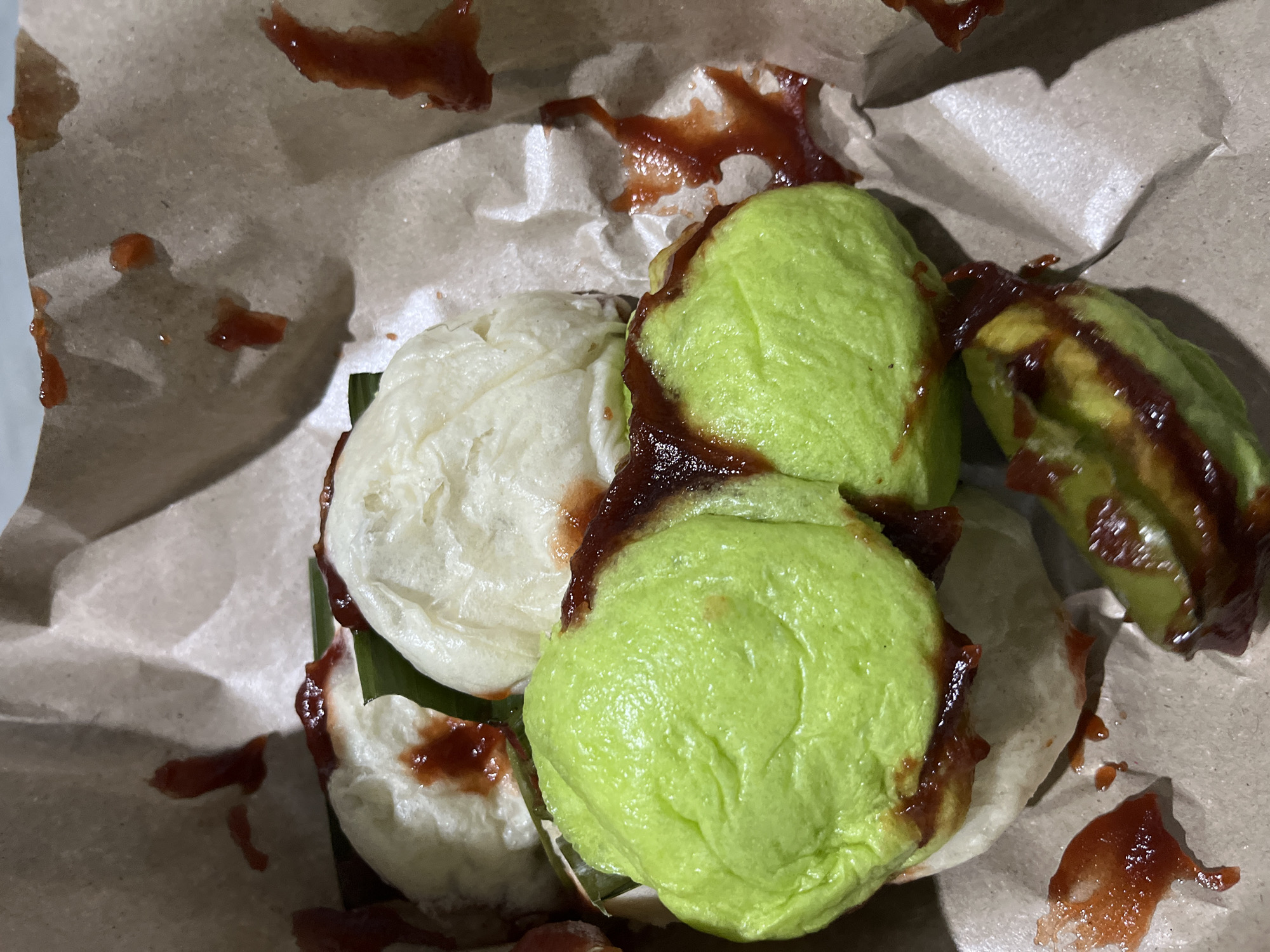 A close-up overhead shot shows a serving of steamed buns, possibly "bakpao," presented in crumpled brown paper packaging. Some of the buns are white, while others are a vibrant green, suggesting different flavors or fillings. Both types of buns are topped with a dark red sauce, likely chili sauce or ketchup, which has also splattered onto the surrounding paper. A few pieces of green leaf, possibly banana leaf, are visible peeking out from under one of the white buns. The lighting is dim, and the focus is primarily on the food. The image does not depict any specific location in Beuringen, Indonesia, or any people.