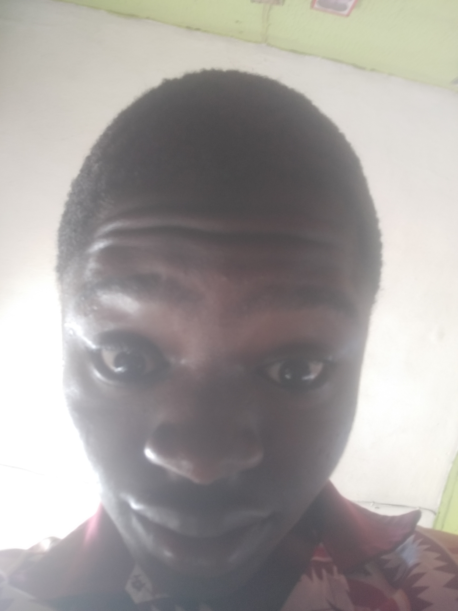 A close-up, slightly high-angle selfie shows a dark-skinned man with short, dark hair. His eyes are wide, and his expression is difficult to discern due to the lighting. He is wearing a collared shirt with a print, appearing to be red and white. The background is blurry, but hints of a light green wall and white surfaces are visible. There is no definitive evidence to place the subject specifically in Shangisha, Nigeria.