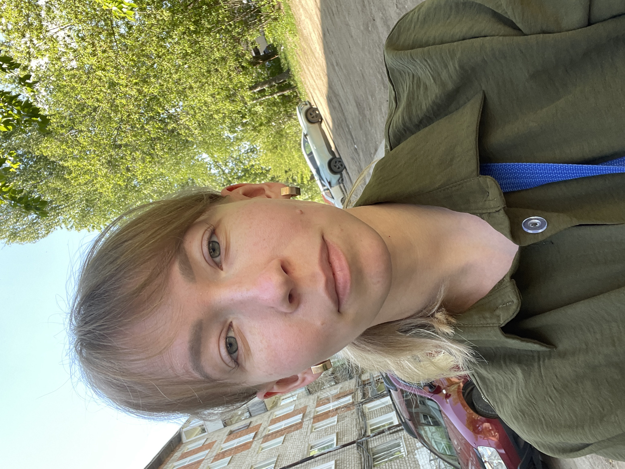The image is a selfie taken by a fair-skinned person with blonde hair and gray eyes. The person is wearing a green collared shirt with a blue strap visible beneath the collar. The background includes a building with multiple windows, trees with green foliage, and a car. The setting appears to be an urban environment in Perm, Russia.