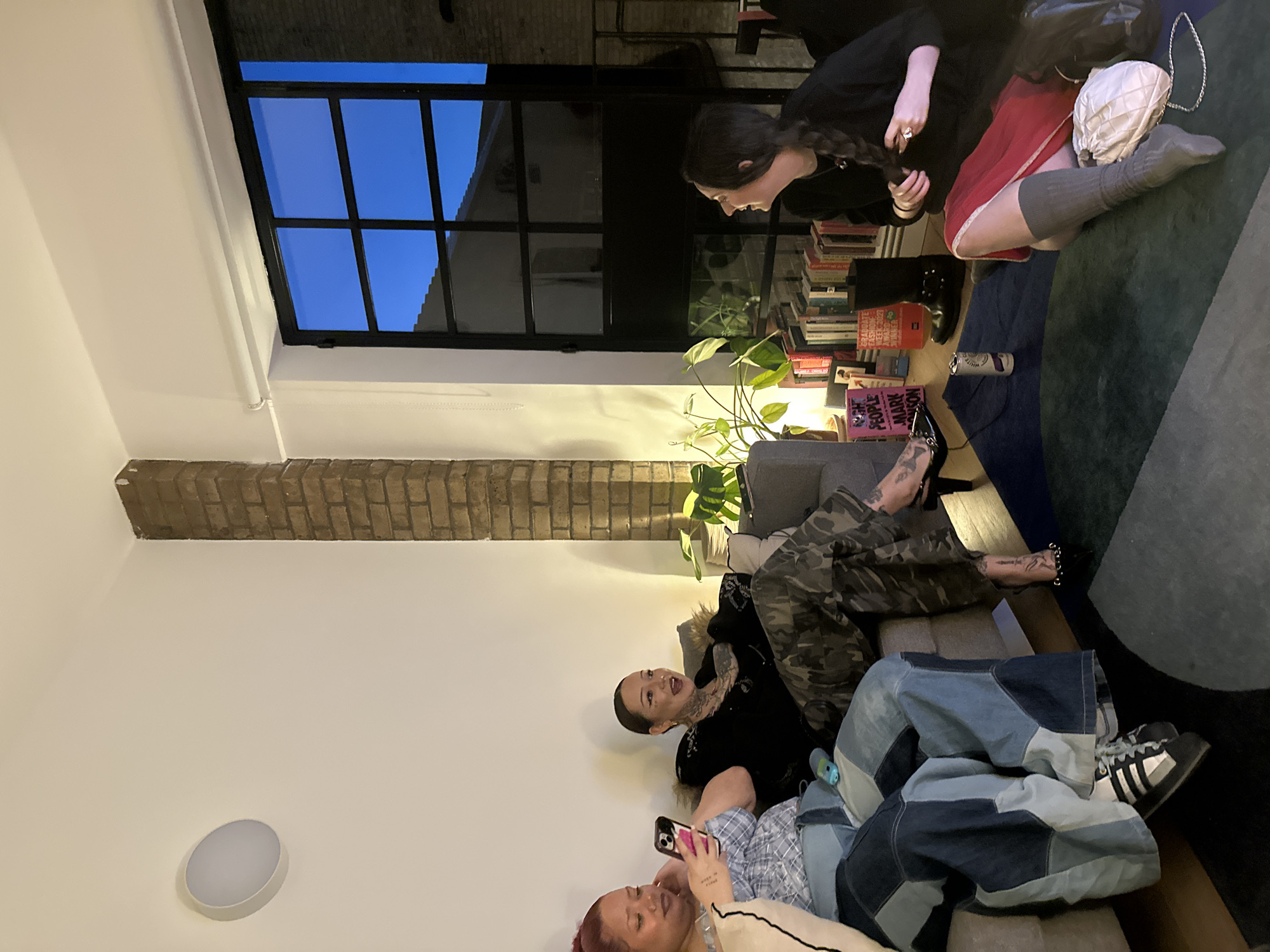 The image shows a gathering of people indoors. Three individuals are reclining on a sofa, with one looking at a phone. Another person is seated on the floor with a book collection nearby, looking at someone else. The setting appears to be a residential or apartment space, featuring a large window showing a cityscape, possibly in the evening. The architectural details suggest a location in London, United Kingdom. There's also a potted plant and a lighting fixture visible. The overall ambiance suggests a casual, social occasion.