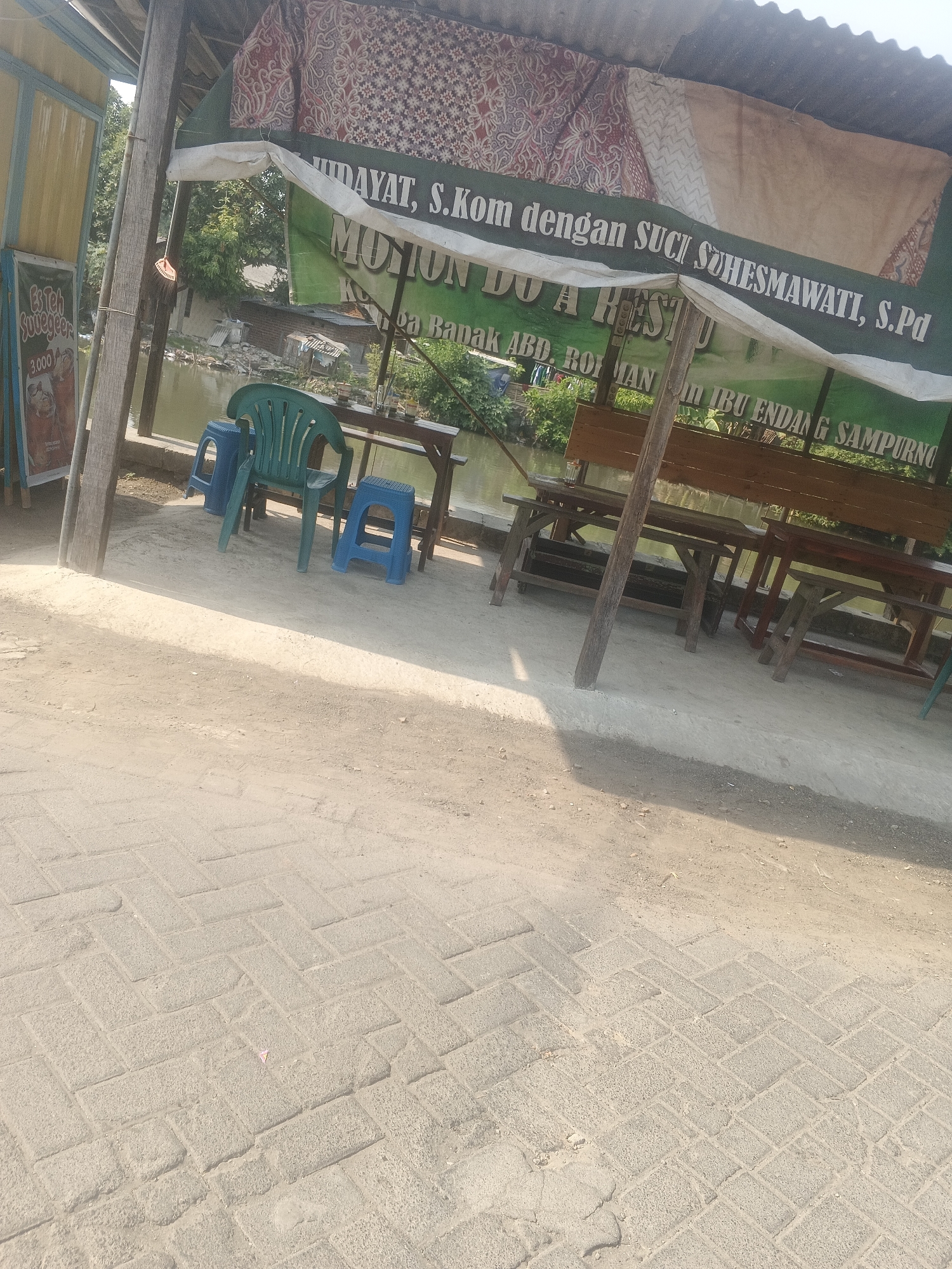 This is a medium shot of a roadside stall in Krian, Indonesia. The stall has a green tarp for a roof, with a traditional batik fabric hanging at the front. A sign in green lettering on the tarp reads "MOHON DOA RESTU" and "BAPAK ABD. ROHMAN". To the left of the stall is a poster for "Es Teh Surabaya" priced at 3,000. The stall is equipped with wooden tables and chairs, some of which are green plastic. A body of water, likely a river or canal, is visible behind the stall. The ground in front of the stall is paved with interlocking bricks. The scene is illuminated by natural sunlight, casting long shadows on the ground.