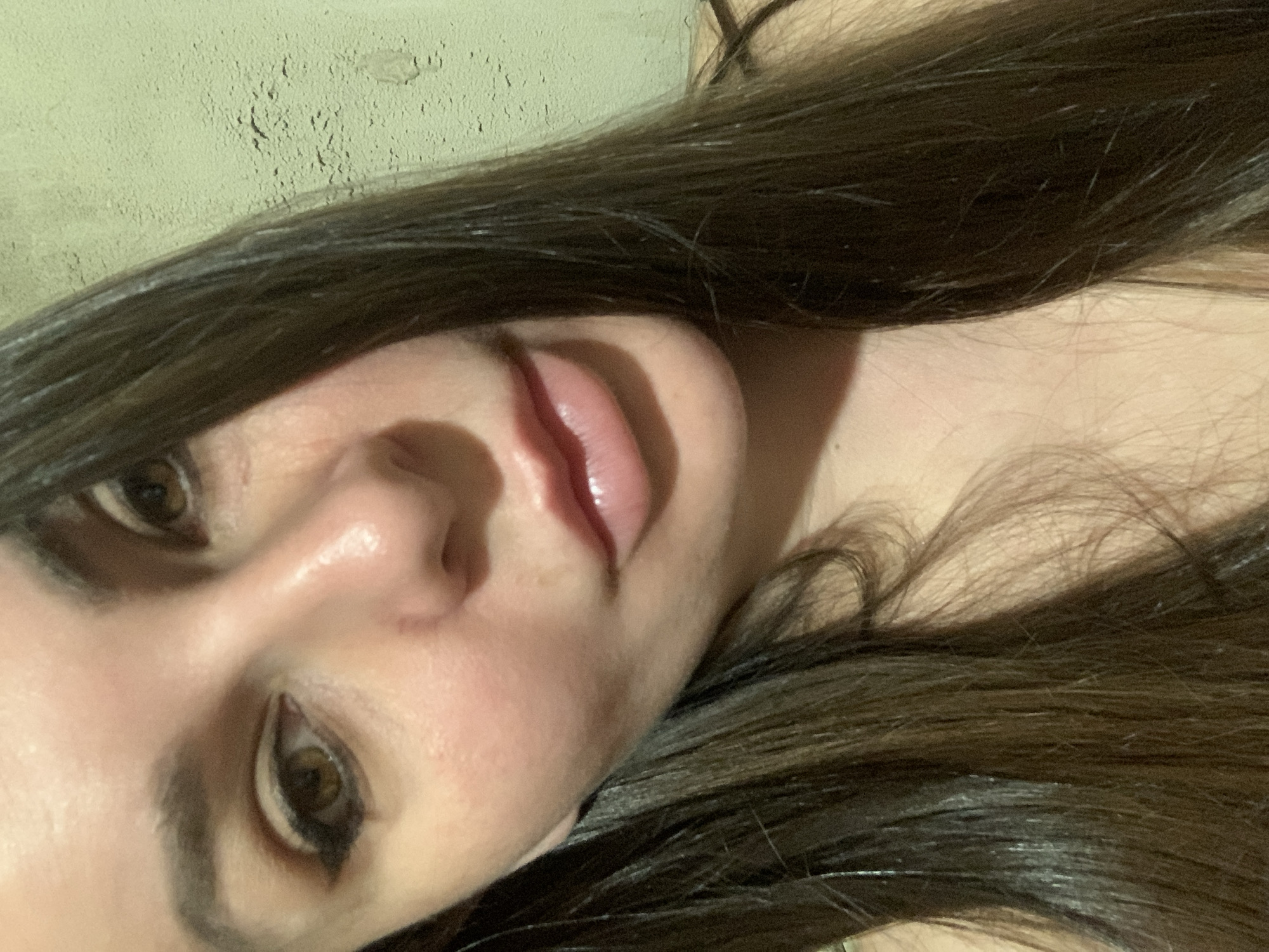 A close-up, overhead shot shows the right side of a woman's face, from her eye to her lips, with her dark brown hair framing her features. The woman's eye is open, and her gaze is directed slightly downward. She has dark eyeliner on her eye and a lighter shade of eyeshadow. Her lips are slightly parted, with a glossy appearance. The background is a textured, pale green wall. The lighting is dim, casting shadows on her face and highlighting her skin. The image is a selfie, as suggested by the close proximity and angle of the camera. The location is not specified beyond "Unknown, Unknown".
