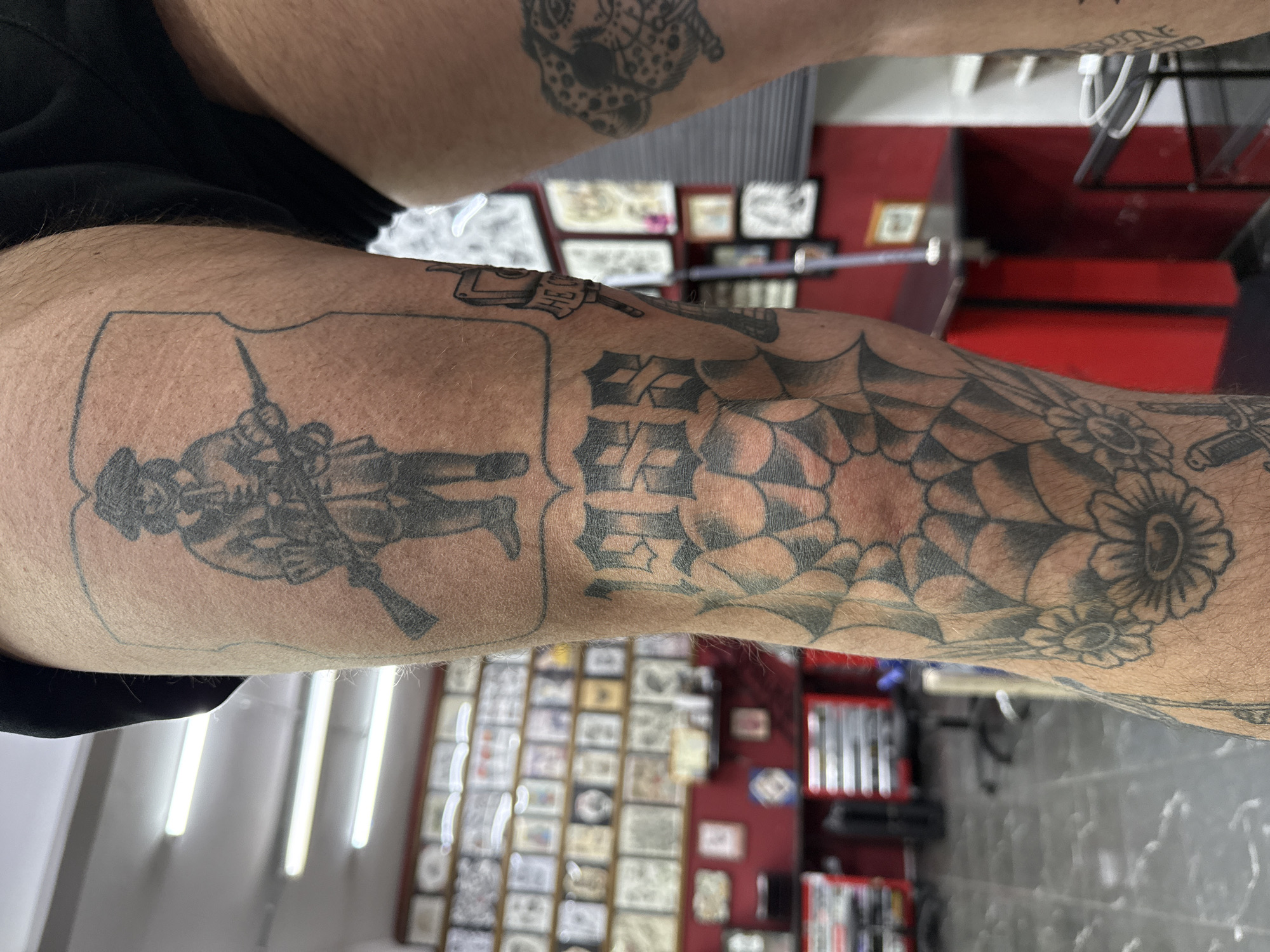 The image shows an arm with multiple tattoos, including a figure with a rifle inside a shield, lettering, a spiderweb with flowers, and other indistinct designs. The arm is light-skinned and hairy. The background appears to be a tattoo parlor, featuring framed artwork on the walls, shelves with items, and various equipment. The location in Moscow, Russia, is not determinable from the image.
