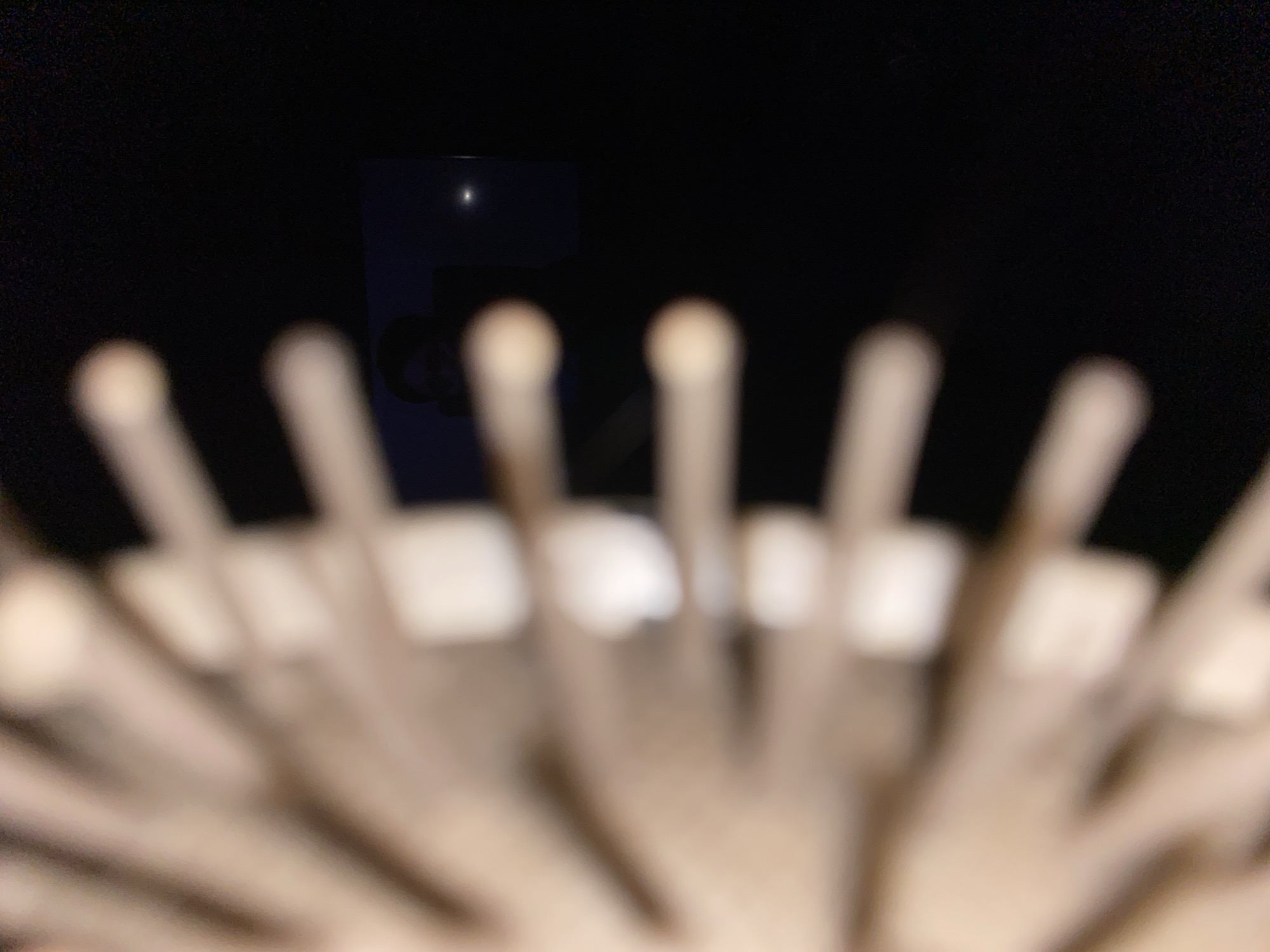The image is a blurred close-up of multiple drumsticks arranged in a rack or holder. The background is dark, with a blurred silhouette of what appears to be a building or structure visible. A small bright point of light is visible near the top of the building, perhaps a streetlight or a reflection. The foreground drumsticks are out of focus, but their cylindrical shape and round tips are discernible. No people are visible. The image is likely taken indoors, given the darkness and the proximity to the drumsticks. The location, Chaikovsky, Russia, cannot be confirmed from the image alone. There is no information to indicate the action, interactions, or the environment.