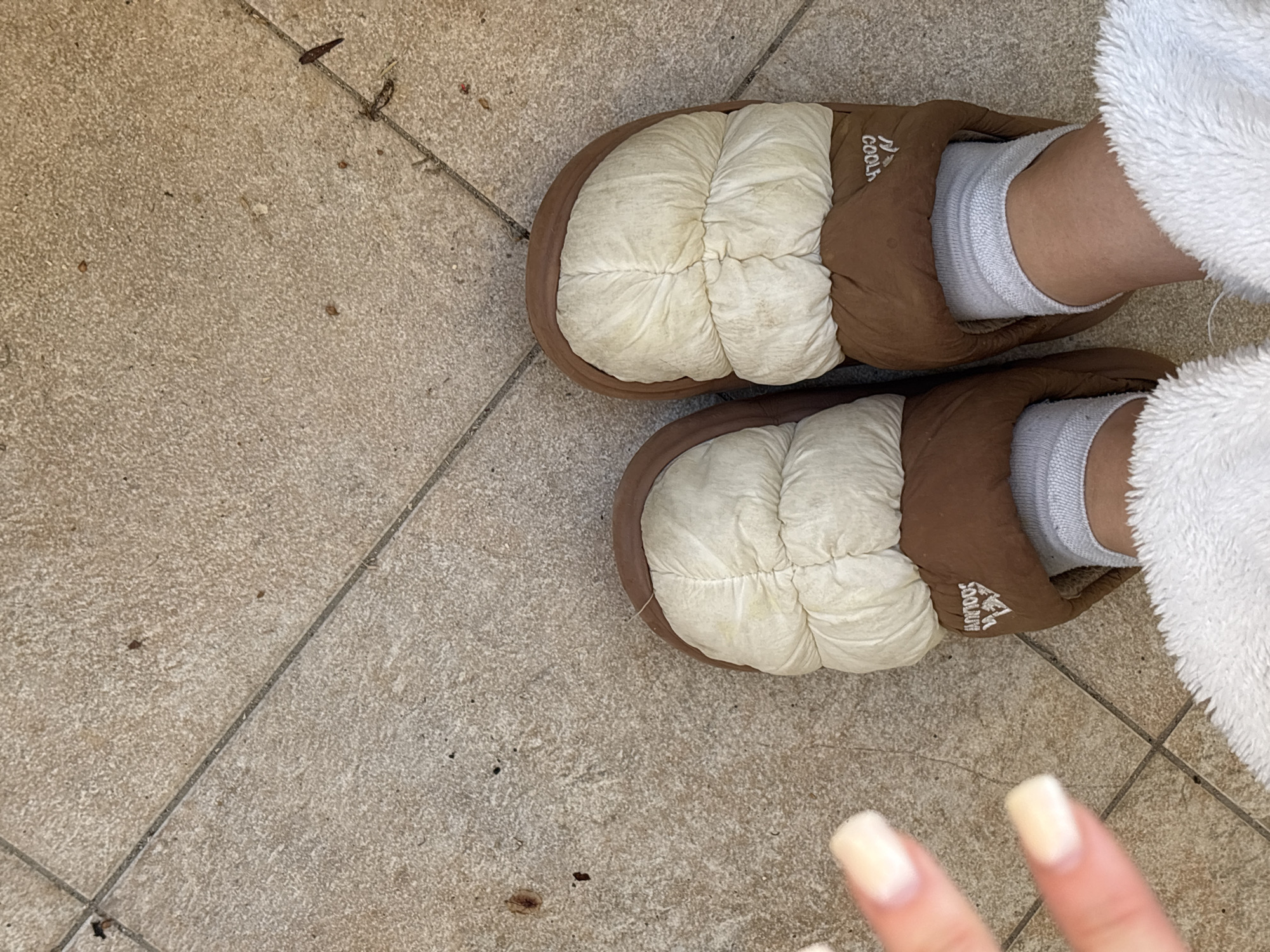 This overhead shot, set in Bečići, Montenegro, captures a person's lower legs and feet dressed in casual attire. The individual is wearing white ankle socks and a pair of two-toned slippers. The slippers feature a brown exterior and an off-white, quilted upper section. Visible on the side of the right slipper is the brand name "COOLOUT" in white lettering, alongside a small mountain logo. The person's left leg is partially covered by a fluffy white garment, possibly a robe or blanket. A hand with manicured, light-colored nails is partially visible in the bottom right corner of the frame. The setting is a tiled floor, composed of light beige or tan textured tiles with dark grout lines. Scattered across the tiles are small pieces of debris, such as dirt or dried plant matter, suggesting the location could be an outdoor space like a balcony or terrace, or a high-traffic indoor area. The natural, bright lighting indicates it is daytime. No specific activity is depicted, rather a static pose suggesting relaxation or a brief pause.
