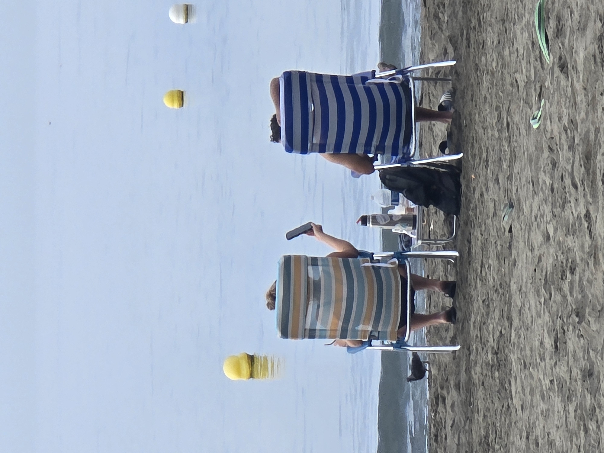 Two people sit in beach chairs on the sand facing the ocean. The person on the left, in a striped chair, holds a phone in their right hand and appears to be looking at it. The person on the right, in a blue and white striped chair, has their head resting on their arm. A small table with bottles and a backpack is between the chairs. A bird stands on the sand to the left of the chairs. Yellow and white buoys are visible in the water. The scene is on a sandy beach, likely in Málaga, Spain, given the presence of beach chairs and the coastal environment.