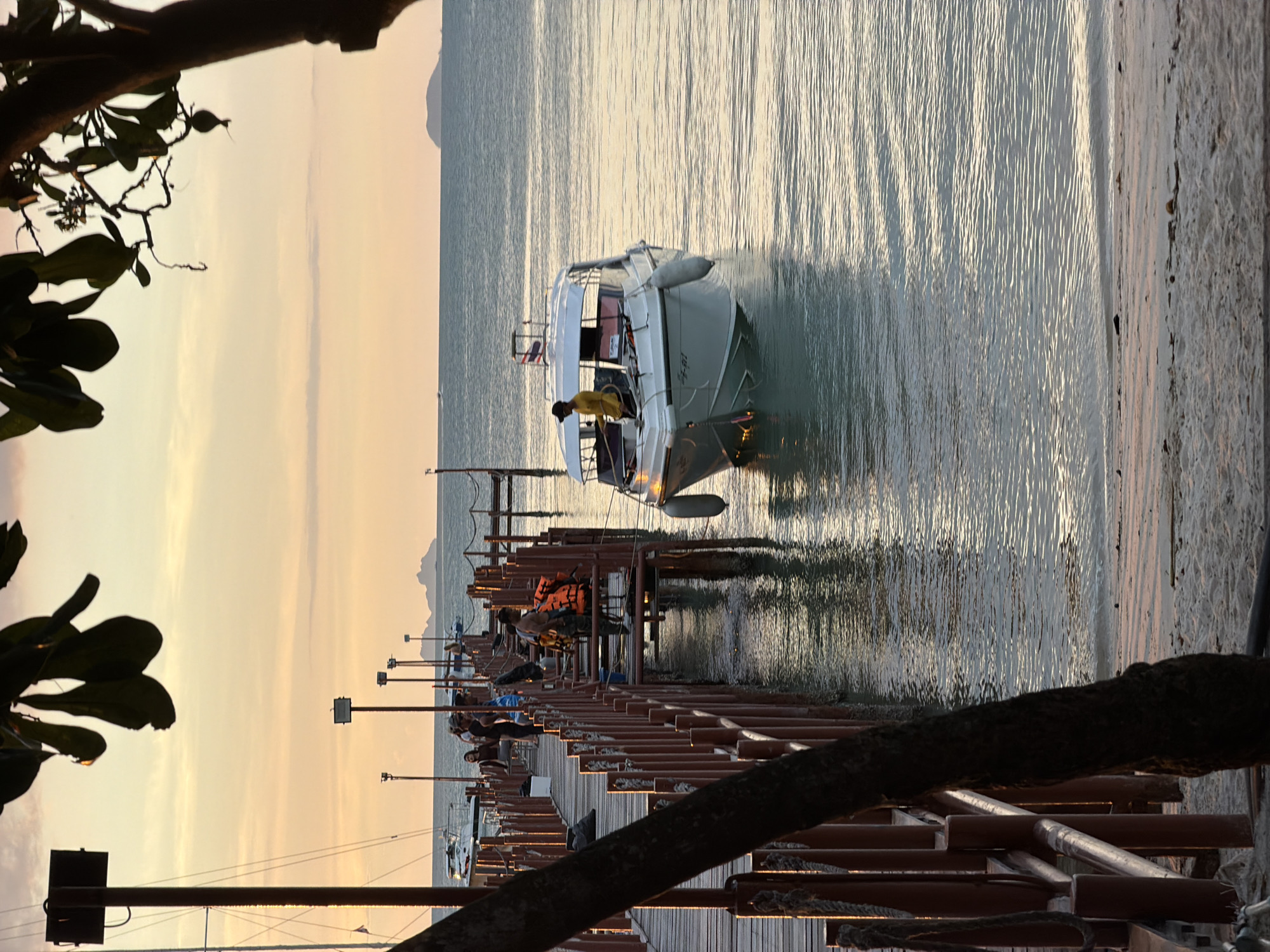 At sunset, a pier extends into calm waters in Baan Bang Rak, Thailand. A white motorboat with a person wearing a yellow shirt and black cap at the helm is docked on the right side of the frame. The boat has a Thai flag on its stern and two white fenders at its bow. A wooden pier, lined with red railings and posts, juts out into the water on the left, with several people visible on it. A tree with dark bark and green leaves is in the foreground on the left, partially obscuring the view of the pier. In the background, a calm sea stretches to the horizon, with distant islands visible. The sky is filled with the warm hues of sunset, with soft clouds.