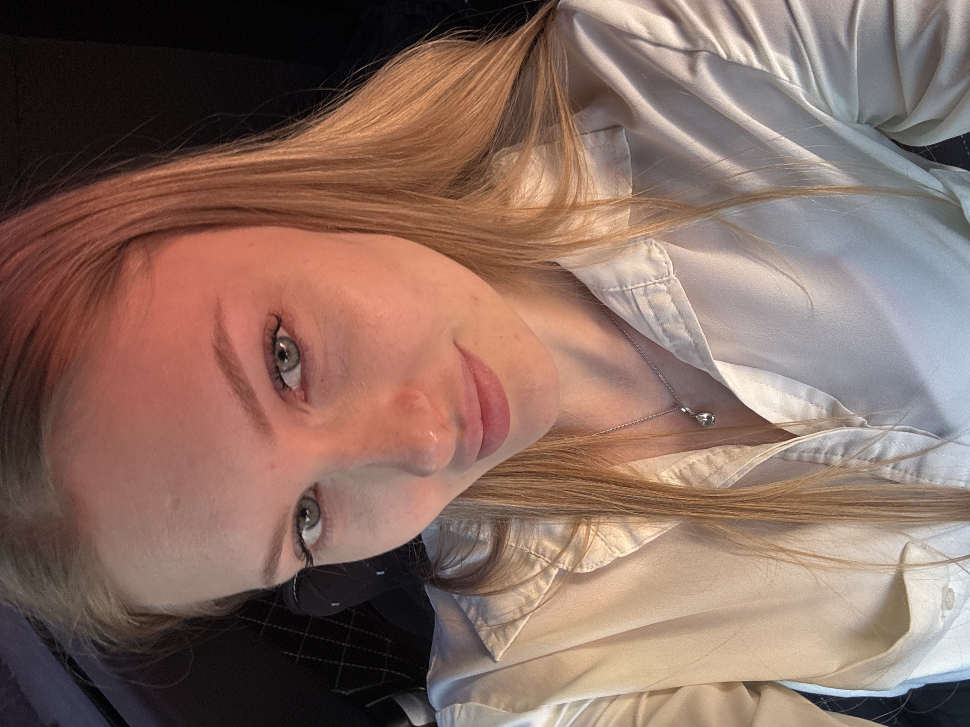 A fair-skinned young woman with light blonde hair is facing the camera. She has light eyes, visible eyelashes, and light eyebrows. She's wearing a white button-down shirt and a delicate silver necklace with a small pendant. The background is dark, suggesting an indoor environment. The image appears to be taken in portrait orientation, possibly as a selfie. There is no information available to suggest a specific location in Perm, Russia.