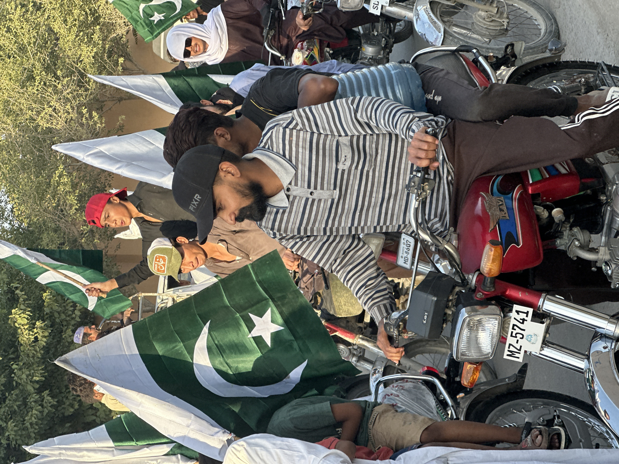 The image shows a group of people, seemingly young men and boys, gathered together, many on motorcycles, and holding Pakistan flags. The motorcycles are parked closely, and some people are resting on them. The scene is likely taking place outdoors in Quetta City Tehsil, Pakistan, though specific location details are not discernable. The setting suggests a gathering or demonstration.