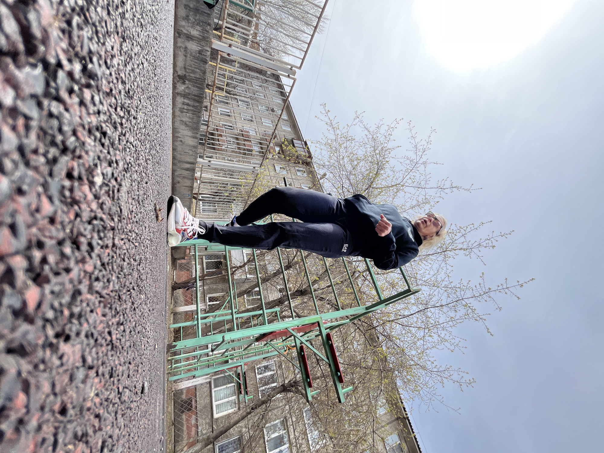 A low-angle photograph shows a woman in athletic wear performing a calisthenics exercise in an urban setting. The woman is wearing a dark-colored tracksuit and light-colored sneakers with red accents. Her blonde hair is short, and she appears to be wearing glasses. She is positioned mid-air, parallel to the ground, with her body aligned horizontally. In the background, a multi-story apartment building with aged, gray facade stands tall. The building features numerous windows and some visible decay. A green metal structure, possibly a jungle gym or playground equipment, is situated between the woman and the building. Bare tree branches with budding leaves frame the sky, which is partly cloudy. The foreground is dominated by a rough, dark-colored asphalt surface. The location is likely an urban area in Krasnoyarsk, Russia, given the style of the architecture. The overall impression is one of someone exercising in a public space within a densely populated residential area.