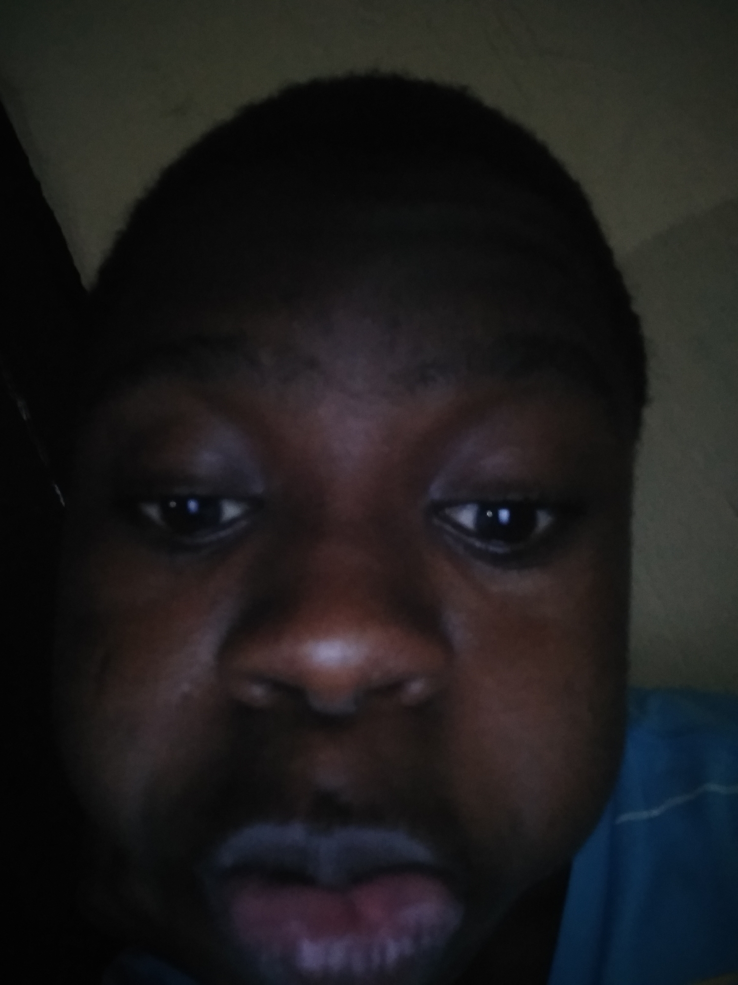 A close-up selfie of a young Black male. He has dark eyes, short hair, and his lips are slightly parted. The background is dimly lit and appears to be indoors. The person is possibly located in Shangisha, Nigeria, based on the prompt request.