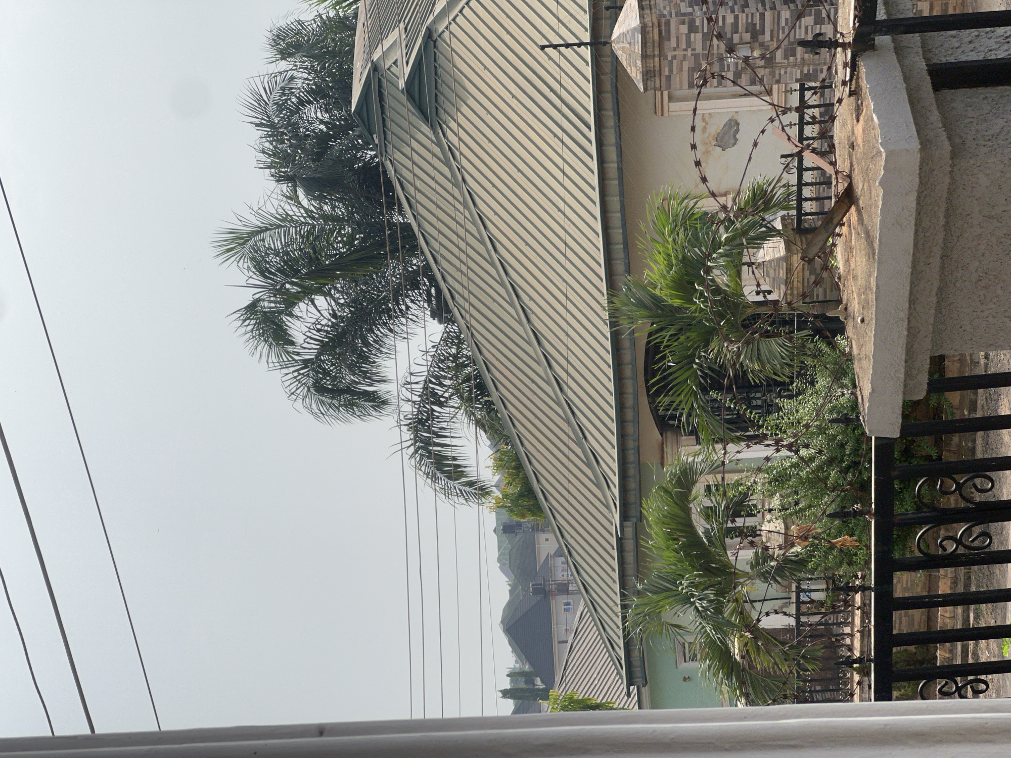 The image shows a view from a higher vantage point, likely a balcony or elevated window, looking out at a residential area. The foreground features a gray railing. In the middle ground, there's a building with a corrugated metal roof, partially obscured by palm trees. A perimeter wall with a decorative metal fence and barbed wire is visible. In the background, one can see other buildings, indicating a densely populated urban environment. Power lines run across the sky. The sky itself is overcast, suggesting a hazy or humid atmosphere. The details suggest the location is likely in Abuja, Nigeria.