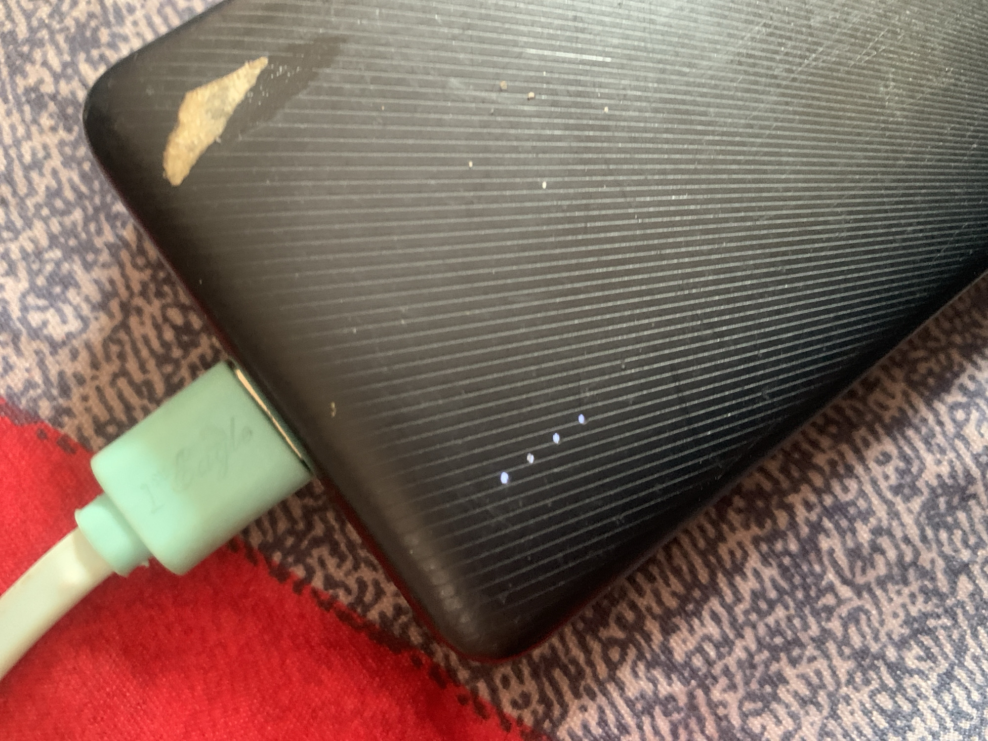 The image shows a close-up of a black power bank plugged into a light green charging cable. The power bank has a ribbed texture and four small white LED lights are illuminated, indicating its charge level. A small, light-colored stain is visible on the upper right corner of the power bank. The charging cable appears to be connected to a device not fully visible in the frame, positioned on a red fabric. The power bank and cable are resting on a patterned surface with shades of brown and beige. The scene appears to be indoors, with no specific details suggesting a location in Jauro Yinu, Nigeria.