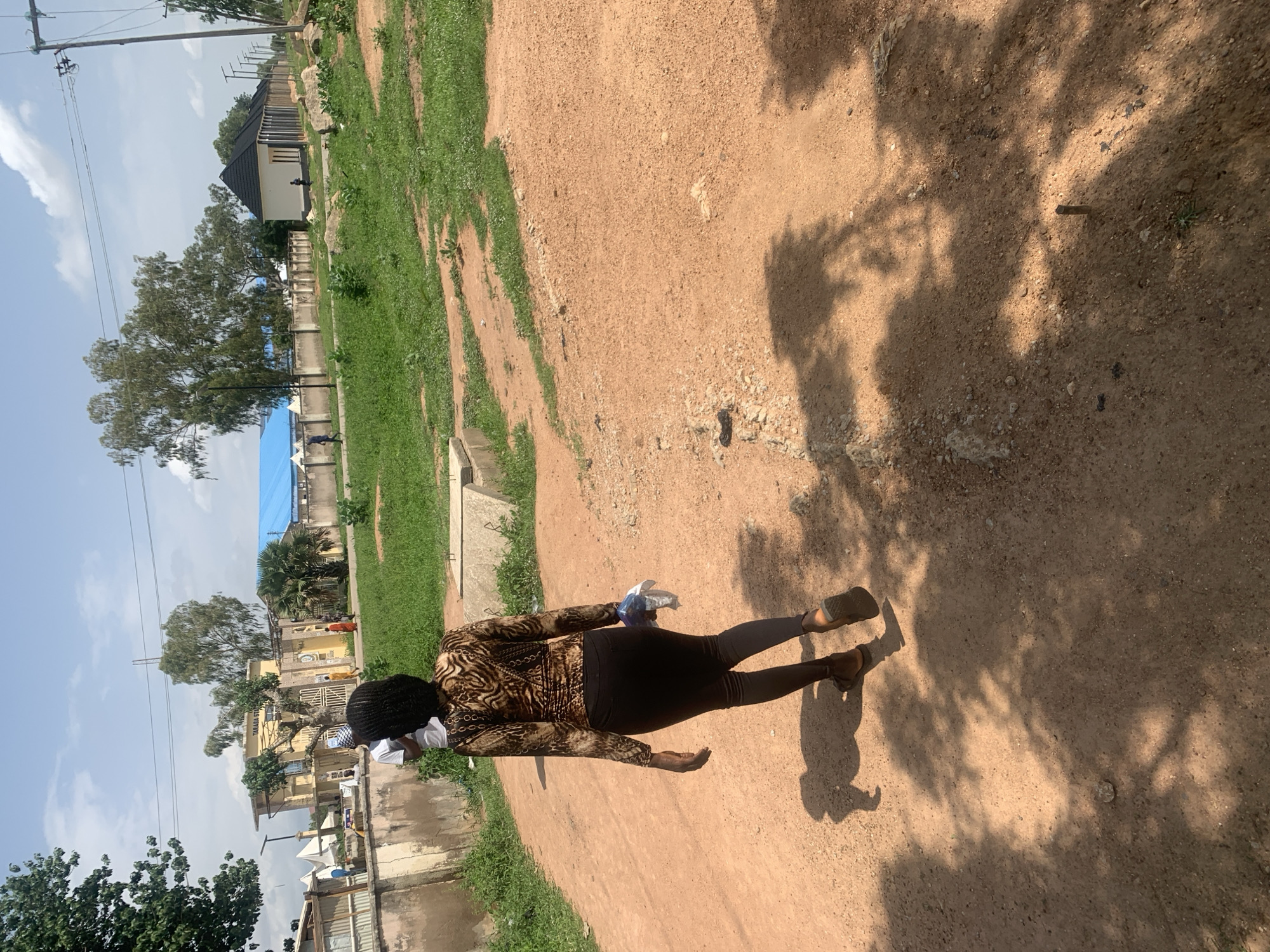 The image shows a woman standing on a dirt path in Jalingo, Nigeria. She is wearing a long-sleeved, patterned top and dark leggings, and is holding a clear plastic bag with some white items inside. Her back is to the camera, and she appears to be looking downwards.
The setting is outdoors, with a mixture of sparse green grass and dry, sandy soil. In the background, there are buildings with corrugated metal roofs and walls, typical of residential areas in Nigeria. A large tree with dark green foliage is visible, and a blue tarp is stretched over something in the distance. The sky is mostly clear with some white clouds. Sunlight casts shadows on the ground, indicating it is daytime. The overall impression is one of a simple, everyday scene in a Nigerian town. There is no visible text in the image.