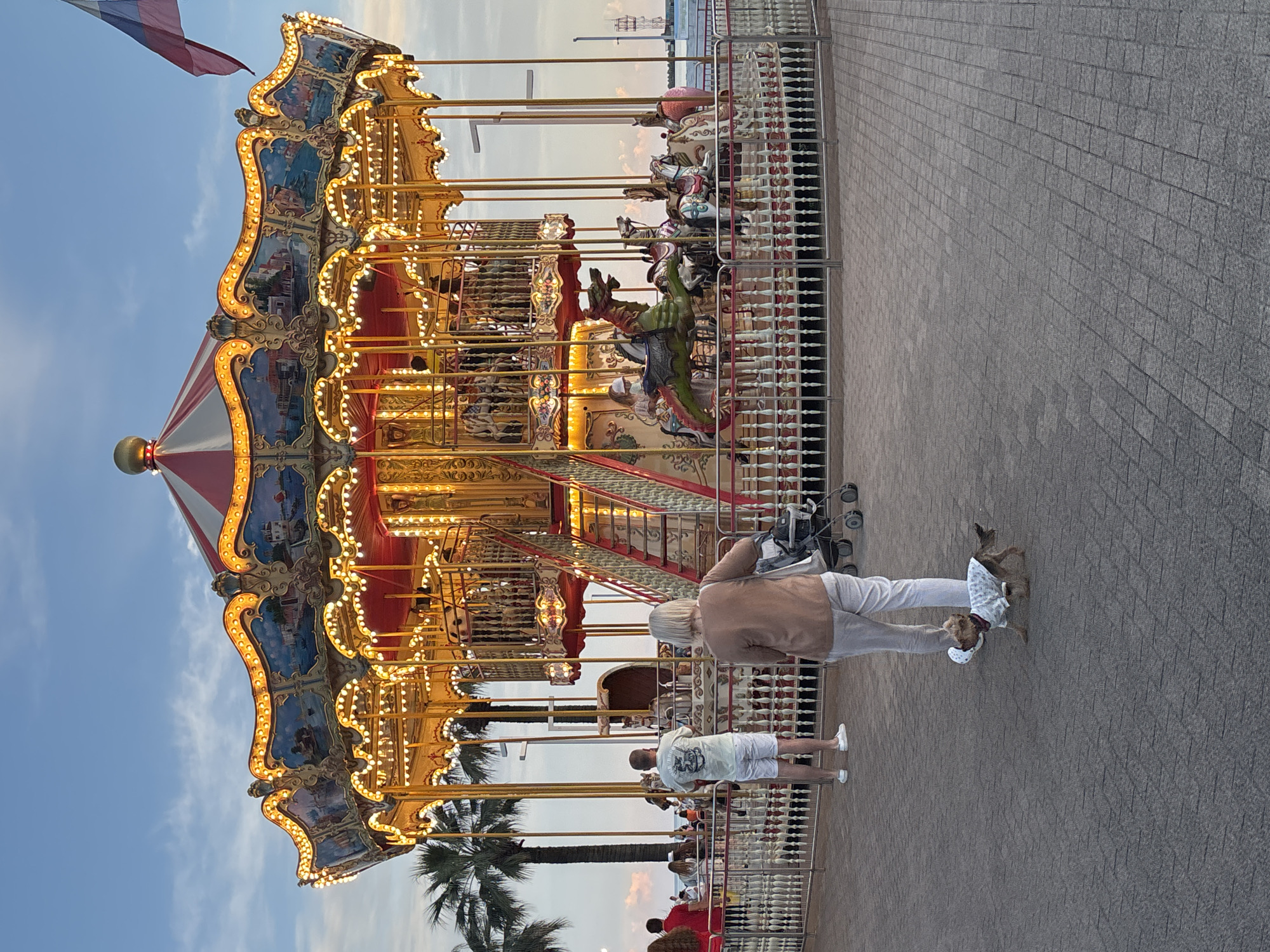 This is a medium shot of a carousel at dusk in Sochi, Russia. The carousel is brightly lit with golden lights and has a red and white striped roof. It is decorated with ornate gold trim and painted scenes of buildings and landscapes. Several carousel horses and a dragon are visible. In the foreground, a woman with blonde hair walks away from the camera, pulling a stroller and holding a leash attached to a small dog wearing a patterned shirt. Behind her, a man in a light-colored t-shirt and white shorts stands facing the carousel. A fence surrounds the carousel, and beyond that, palm trees and the sky are visible. A Russian flag flies in the upper right corner. The ground is paved with interlocking gray bricks.