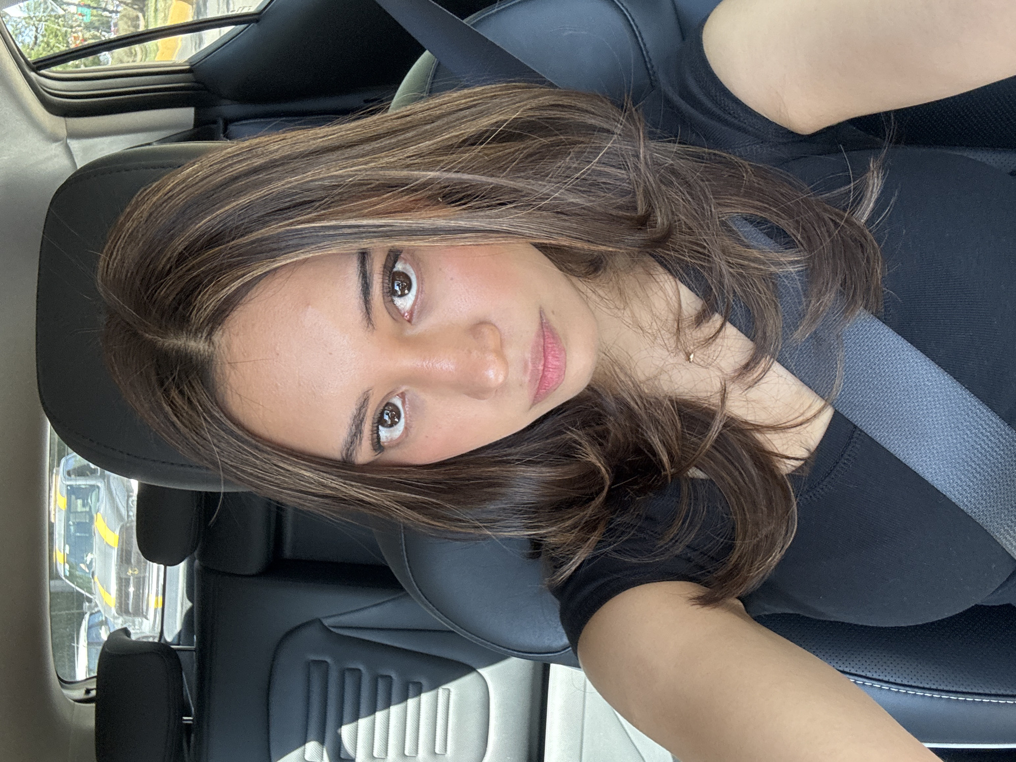 A woman is sitting in the driver's seat of a car, wearing a black shirt and a seatbelt. She has brown hair with blonde highlights and brown eyes. The interior of the car is black leather. The exterior, visible through the side window, appears to be a street scene with vehicles, potentially in Puebla, Mexico. No other people are visible. The woman is looking at the camera.