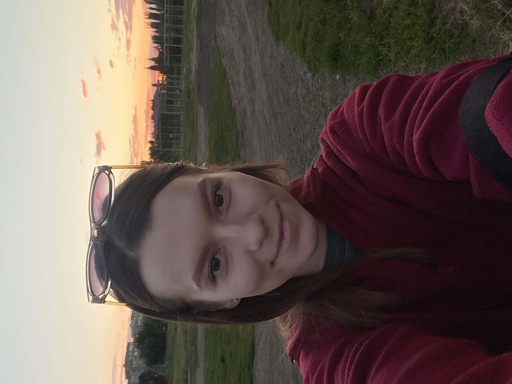 A young woman with dark hair and sunglasses perched on her head takes a selfie outdoors at sunset. She is wearing a burgundy fleece jacket over a dark shirt. The background features a dusty, gravelly area with patches of green grass. In the distance, there are some buildings, trees, and a skeletal structure that appears to be under construction. The sky is a gradient of warm colors, from pale yellow to orange and pink, indicative of a sunset. The image was taken in Kotovo, Russia.