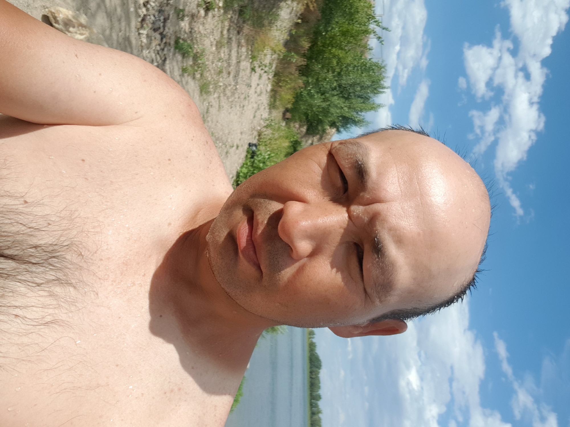 A bald Asian man with dark hair on the sides of his head is shown from the chest up. He has tanned skin with small water droplets on his face, chest, and shoulders. The man is outdoors in what appears to be a natural setting, with a body of water in the background. The sky is blue with scattered white clouds. The terrain behind the man consists of dry, sandy ground with sparse green vegetation and some bushes. The location is identified as Kurchatov, Kazakhstan.