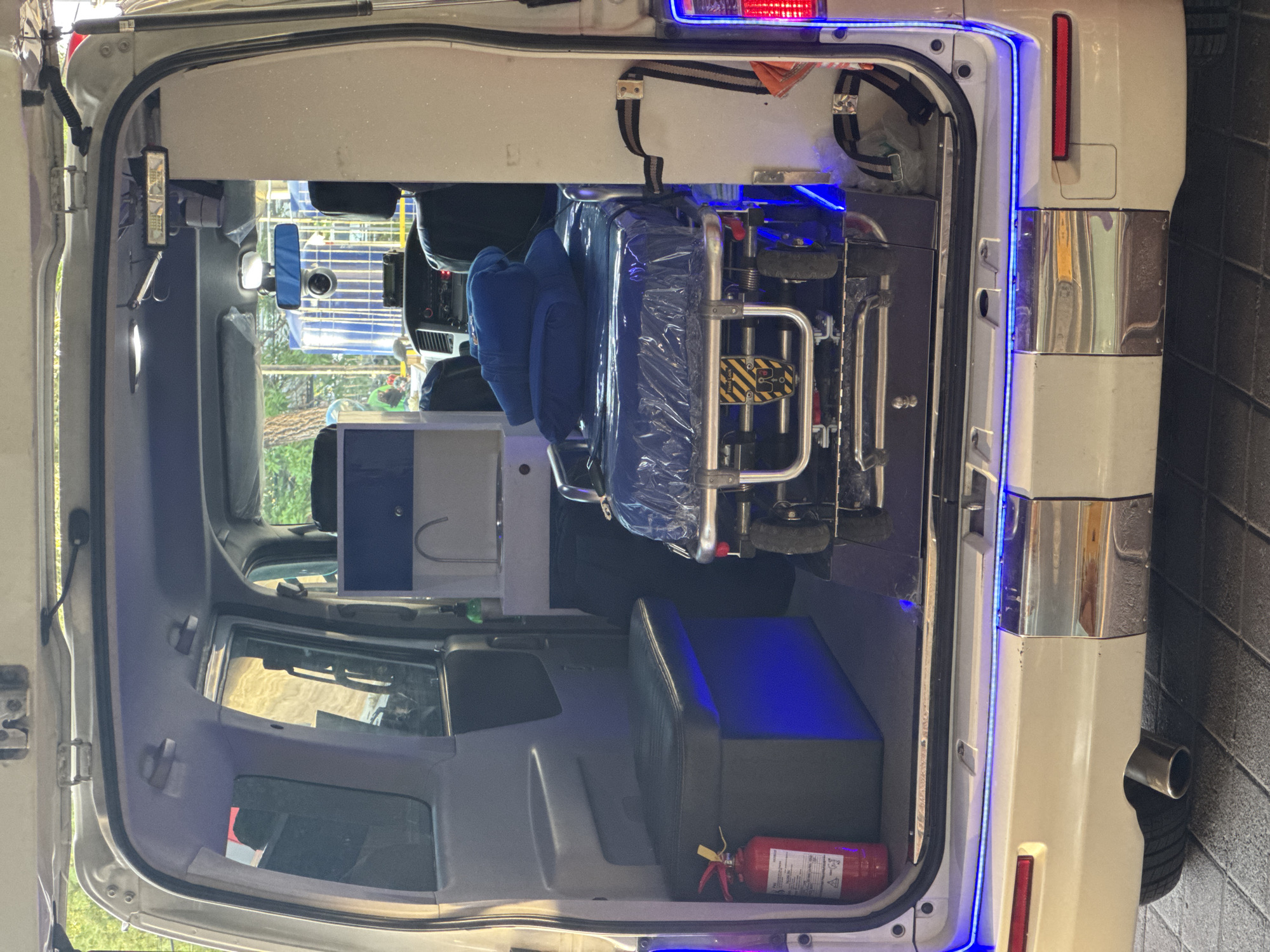 This is a medium shot, taken at a slight upward angle, of the rear interior of an open ambulance, parked outdoors on a paved surface, likely in Kuta, Indonesia, given the provided information. The ambulance is white with blue LED accent lighting visible along the rear bumper and the interior ceiling.
Inside the ambulance, the focus is on the medical equipment. A folded stretcher, covered in plastic wrap and a blue blanket, is positioned centrally. The stretcher has a metallic frame and is equipped with wheels. To the left of the stretcher, a red fire extinguisher is mounted on a grey storage unit. Above the fire extinguisher and storage unit, a blue and white unit with a faucet is visible, suggesting a small sink or water dispenser. To the right of the stretcher, a white panel with straps and medical supplies is partially visible.
In the background, the interior of the ambulance continues, with dark seats and a dashboard area. Through the open rear doors, parts of the outdoor environment are visible, including greenery and what appears to be a building with blue tanks or structures, possibly indicating an industrial or medical facility. No individuals are clearly identifiable within the frame. The overall impression is of an organized and equipped emergency vehicle ready for use.