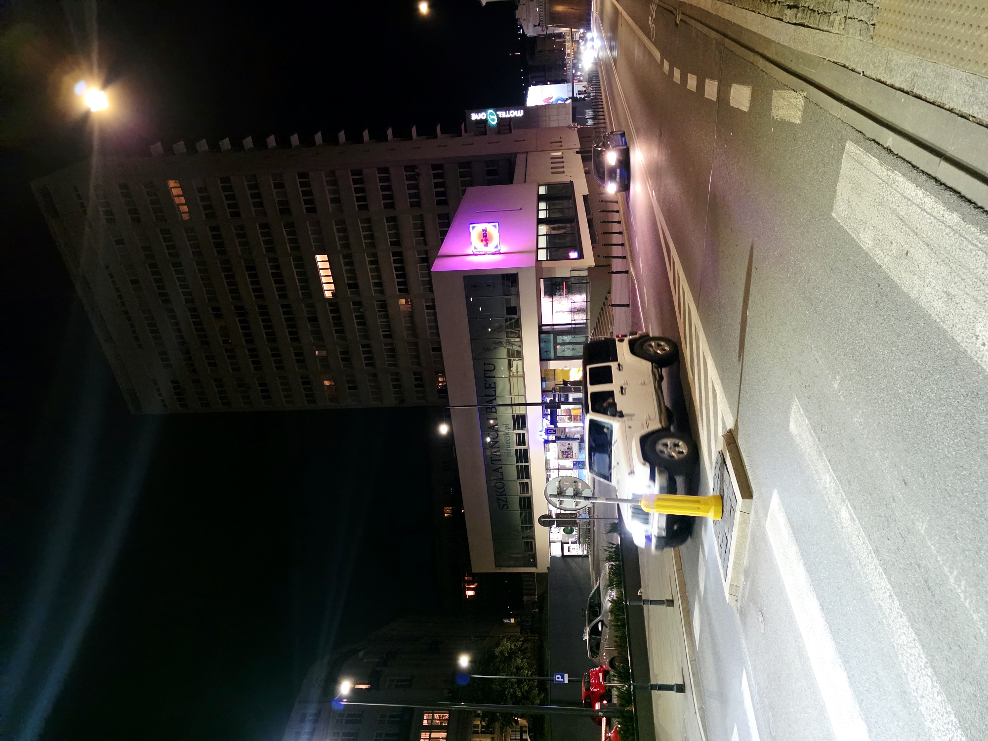 This is a night-time, eye-level, outdoor shot of a city street in Warsaw, Poland. The scene is dominated by a tall, Brutalist-style apartment or office building on the right, with numerous windows, some of which are lit from within. In front of this building, a lower structure has a modern, angular facade with large windows and a prominent pink-lit sign that reads "LIDL." To the left of the frame, a white Jeep Wrangler is stopped at a pedestrian crossing, with its headlights illuminating the road. Behind the Jeep, a silver car and a red car are visible further down the street. The street is paved, with white pedestrian crossing markings and dashed white lines marking traffic lanes. Streetlights and the lights from the buildings illuminate the scene. A tall, modern streetlamp is visible on the left side of the image. The sky is dark.