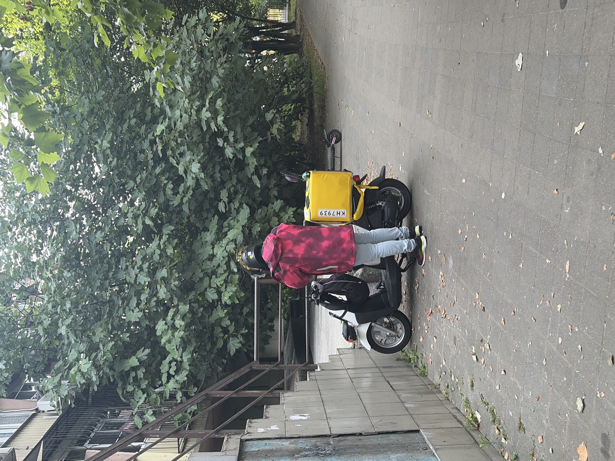 A person wearing a tie-dye jacket and light blue jeans stands next to a white scooter with a yellow delivery box attached. The scooter is parked on a paved walkway lined with trees. A stairway with a metal railing is visible to the left of the scooter, leading up to a building with barred windows. The walkway is covered in scattered fallen leaves. The scene appears to be in an urban outdoor setting.